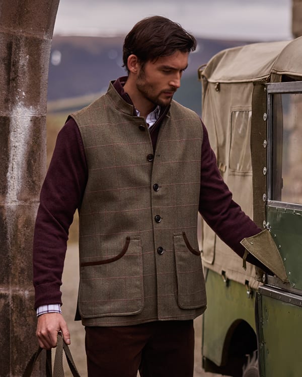 A man wearing a brown tweed waistcoat  over a dark burgundy sweater, standing outdoors next to a vintage green vehicle.