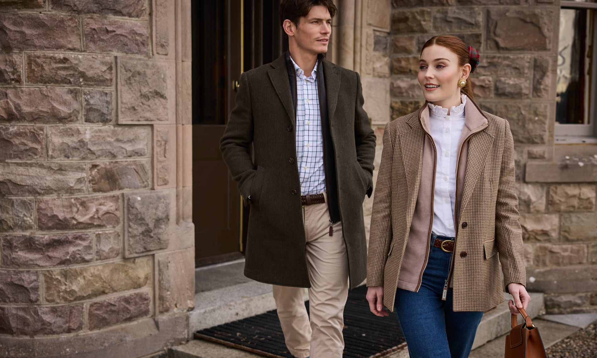 A man and a woman walking out of a stone building. The man wears a dark green tweed jacket over a blue and white check shirt and beige trousers.