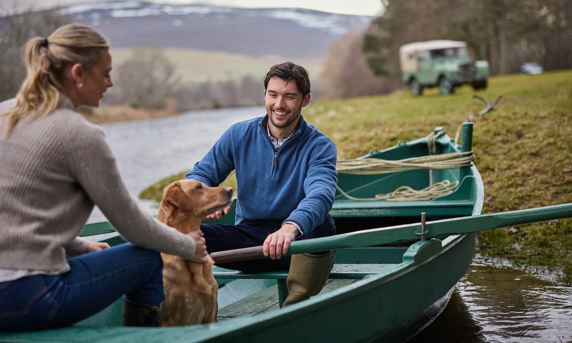 A man and a woman sitting in a green rowboat on a lake, with a  dog beside them and a grassy, mountainous outdoor setting in the background.