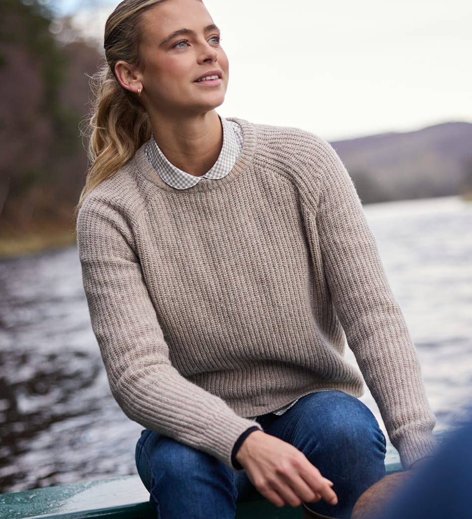 A woman sitting outdoors by a body of water wearing a beige ribbed knit jumper over a gingham shirt and blue jeans.