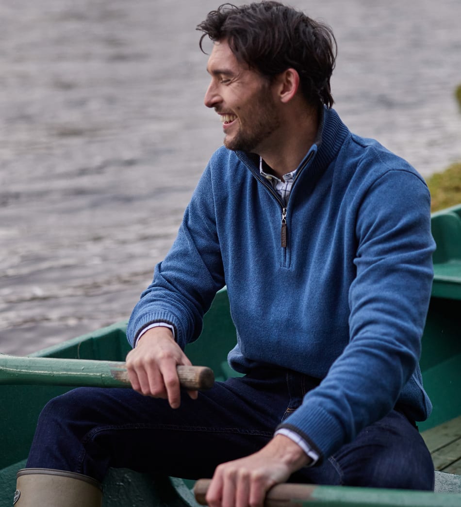 A man wearing a blue quarter-zip knit jumper sits in a green boat on the water, holding oars.