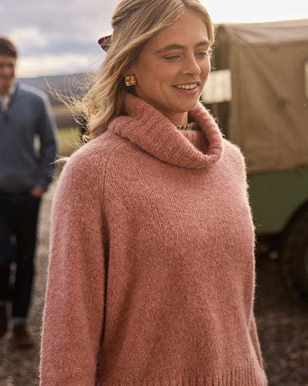 A woman wearing a dusty pink cowl-neck knit jumper, standing outdoors with a blurred man and a vehicle in the background.