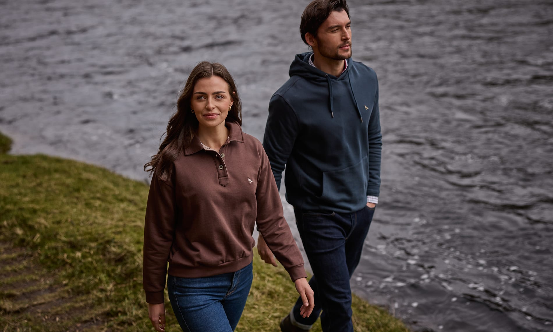 A man and a woman walking outdoors near a body of water. The woman wears a brown quarter-zip pullover and blue jeans, and the man wears a dark navy pullover hoodie and dark pants.