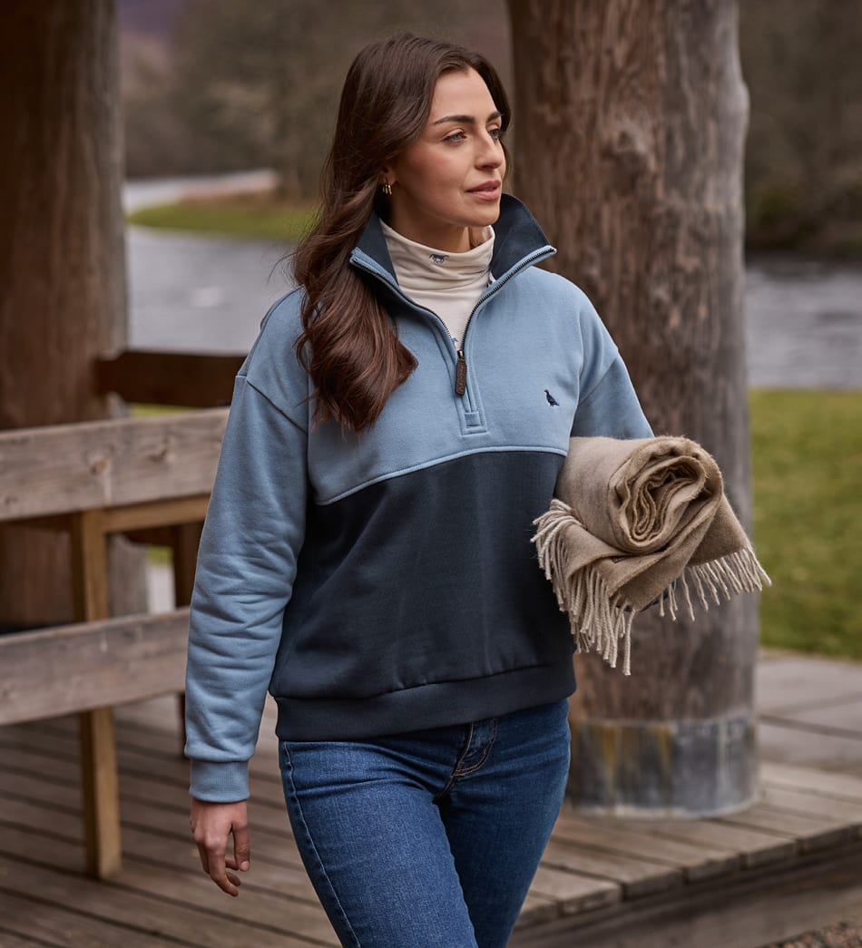 A woman wearing a color-blocked quarter-zip sweatshirt in light blue and dark navy, walking outdoors on a wooden boardwalk while holding a beige fringed throw blanket.