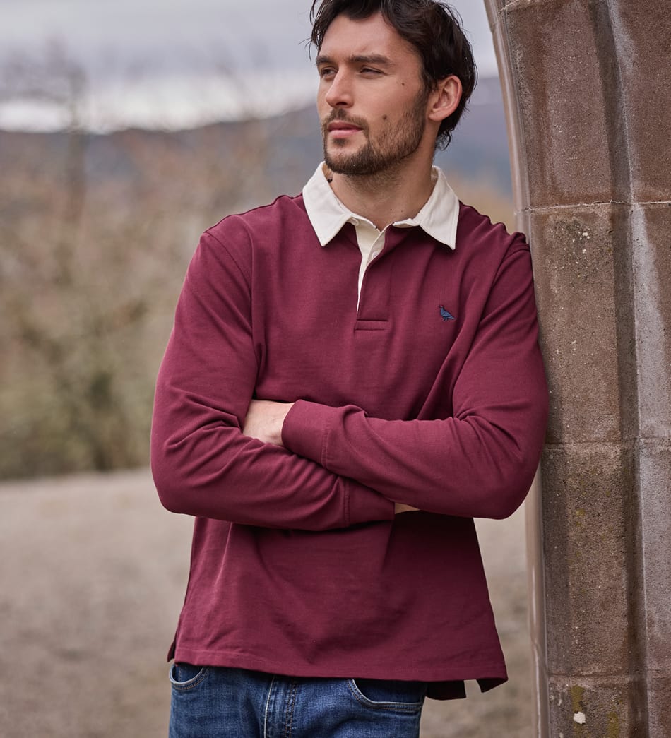 A man wearing a burgundy long-sleeve rugby shirt with a cream collar, leaning against a stone pillar outdoors.