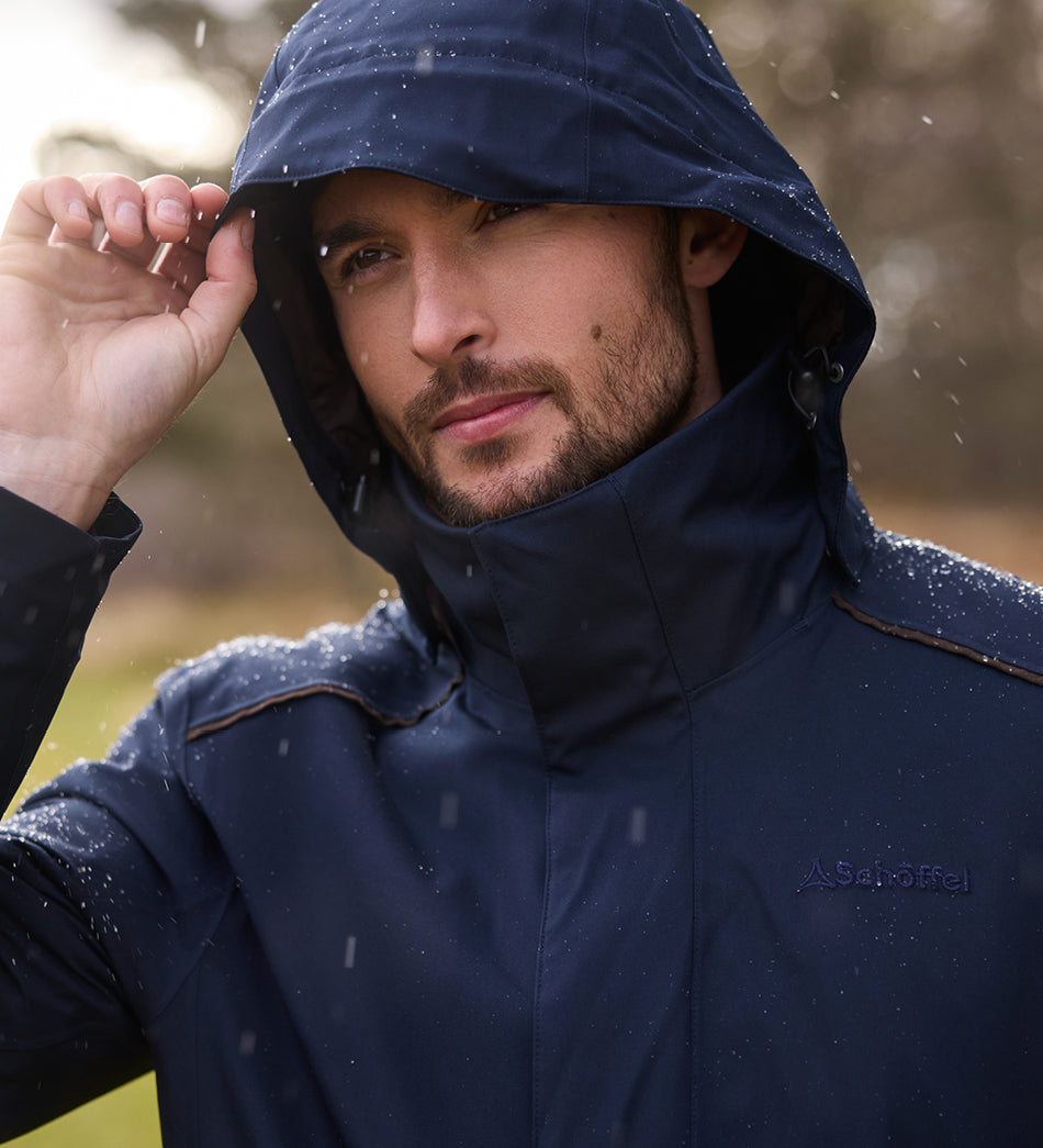 A man wearing a dark navy Schöffel Country hooded jacket in the rain, with water droplets on the fabric and a blurred outdoor background.