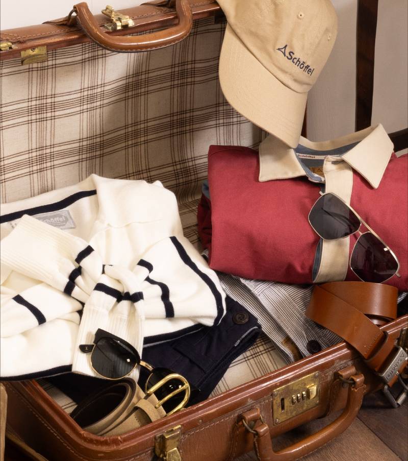 An open vintage brown leather suitcase packed with neatly folded clothing and accessories. Inside are a cream sweater with navy stripes, a red polo shirt with a beige collar, a beige cap, two pairs of sunglasses, a brown leather belt, a tan belt with a gold buckle, navy shorts, and tan loafers. The suitcase is placed on a wooden floor and lined with plaid fabric.