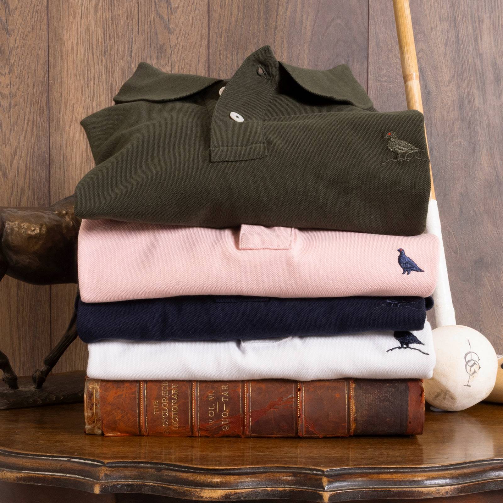 Folded polo shirts in green, pink, navy, and white, each with a Ptarmigan bird detail, resting on vintage leather books.