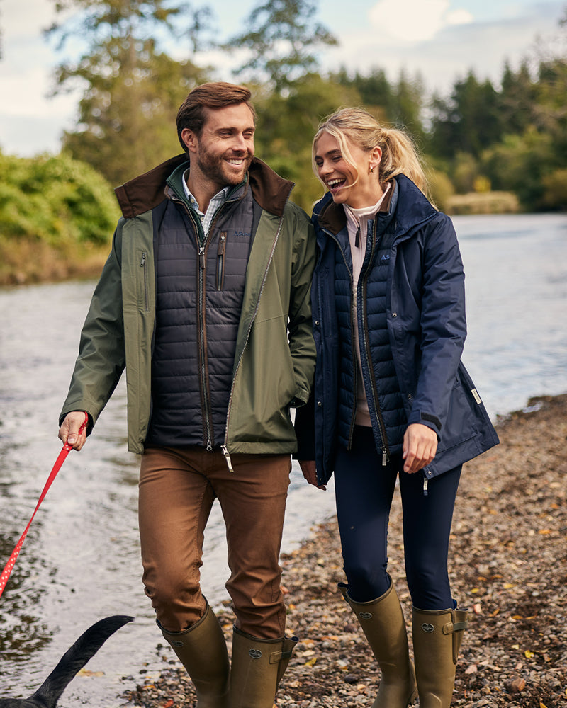 Man and woman walking along a path by a body of water, both wearing insulated jackets.