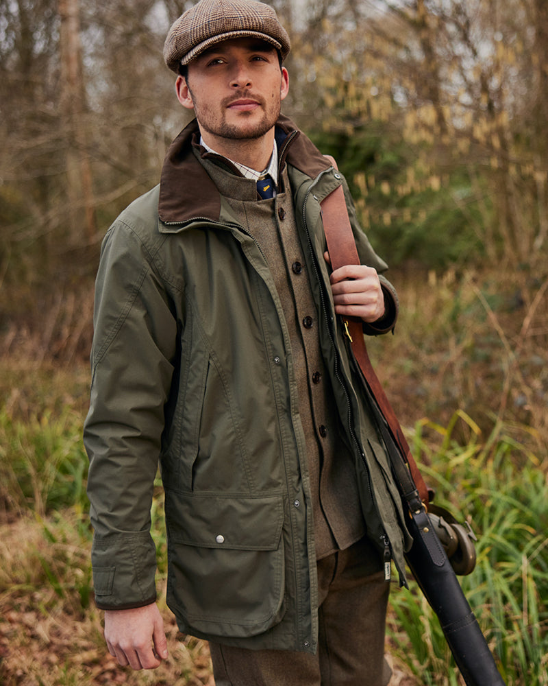 Man in outdoor setting wearing a green jacket and brown cap, holding a gun.