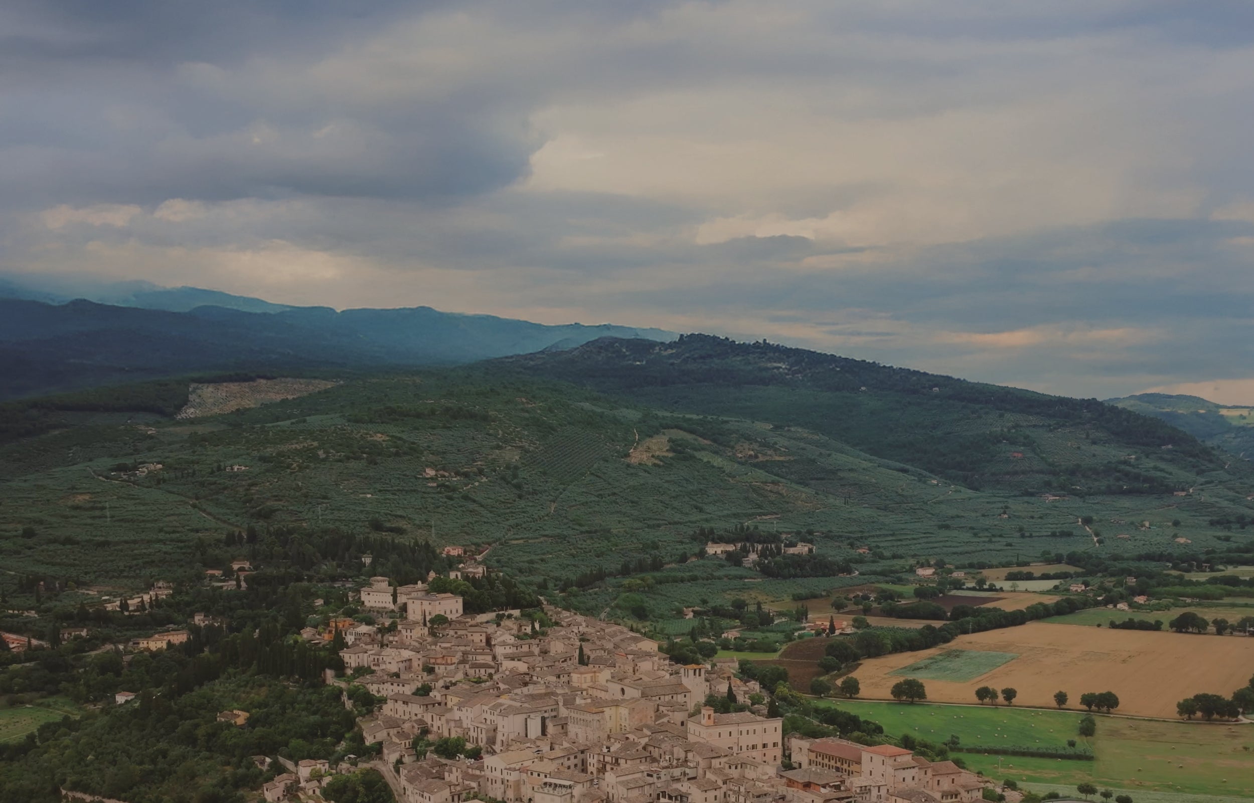 Village nestled in a valley with rolling hills and mountains under a cloudy sky.