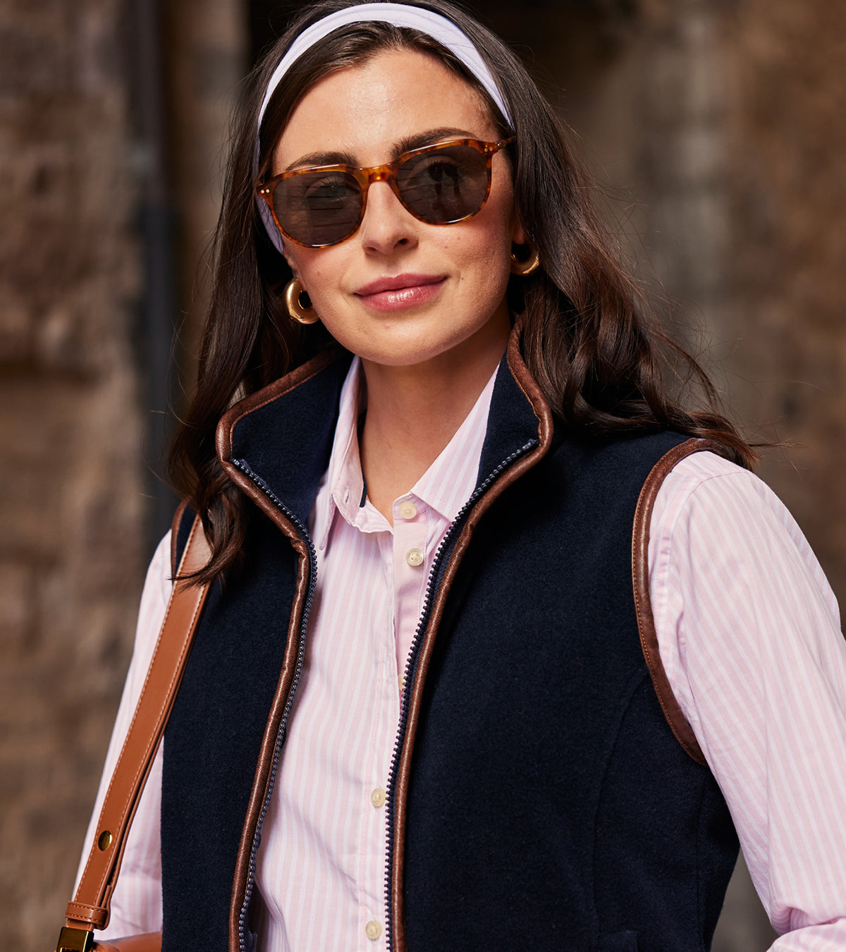 Woman wearing sunglasses, a headband, and a dark gilet over a light shirt against a stone wall.