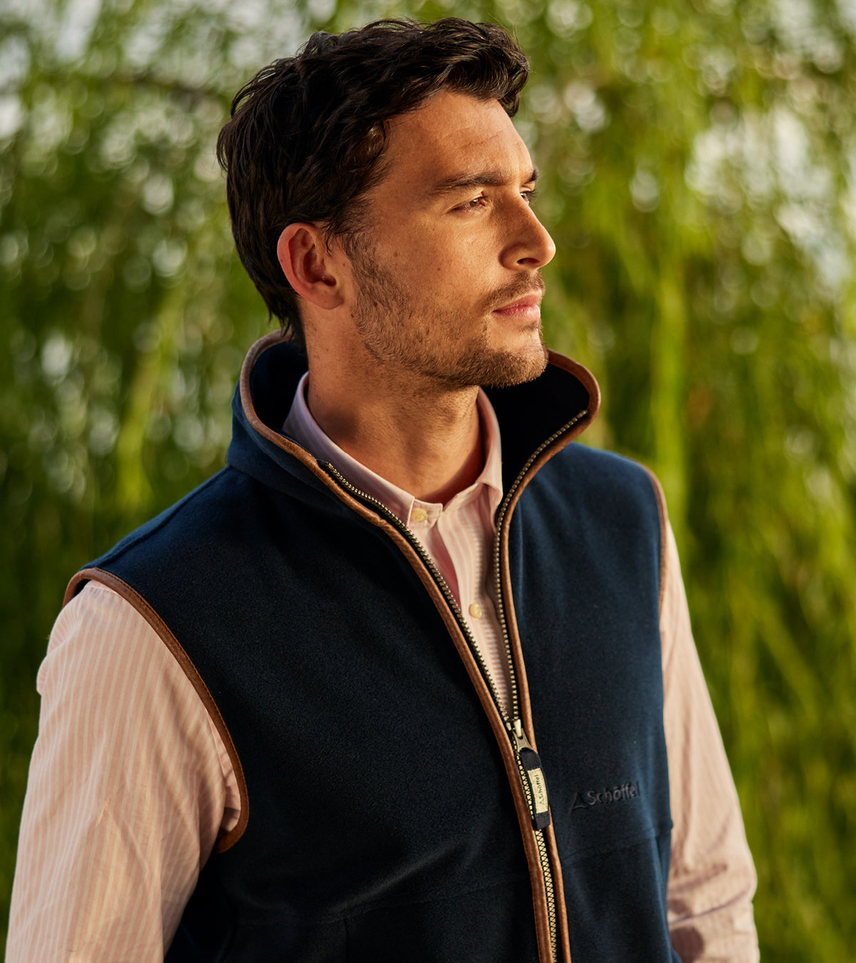 Man wearing a navy gilet over a light pink shirt with a blurred green background