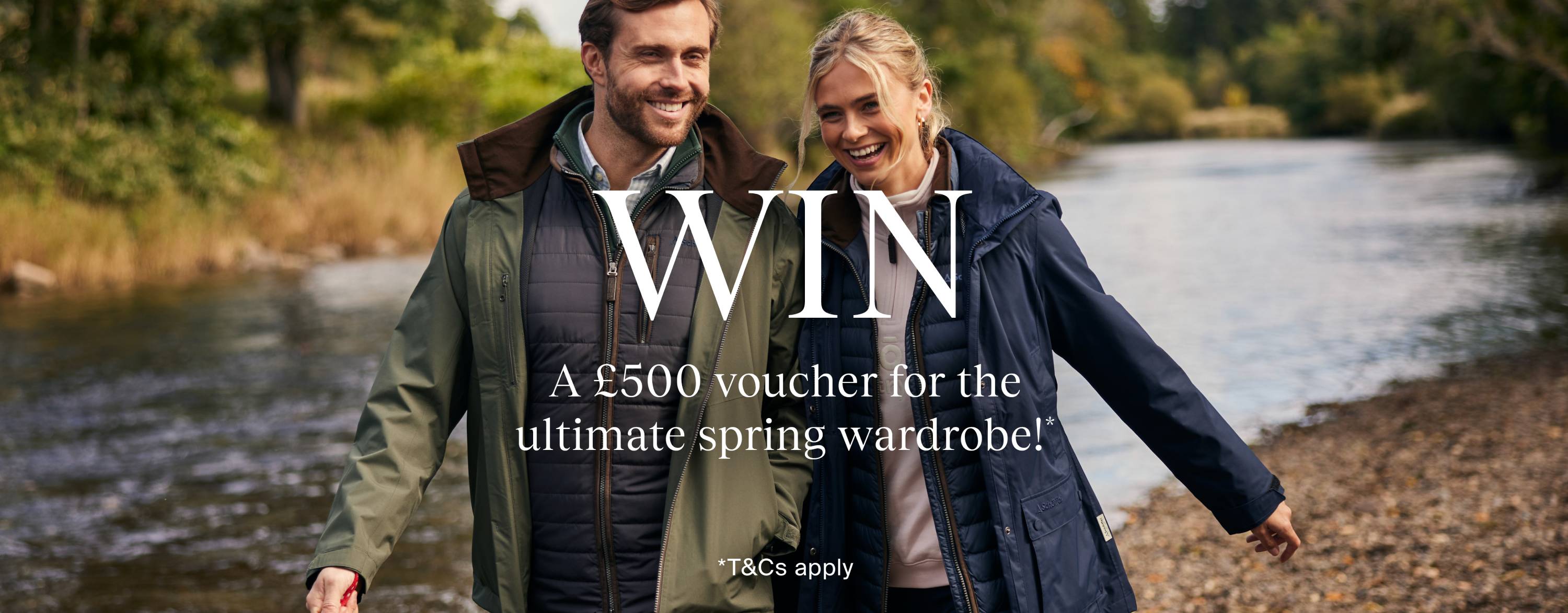 A smiling man and woman walk along the edge of a scenic river, dressed in stylish outdoor clothing. The man is wearing a green waterproof jacket over a navy quilted vest and a checkered shirt, while the woman wears a navy raincoat over a light-coloured sweatshirt. The background features lush green trees and a gently flowing river. Overlaying the image is promotional text in white that reads: 'WIN A £500 voucher for the ultimate spring wardrobe!' with a disclaimer '*T&Cs apply' at the bottom.