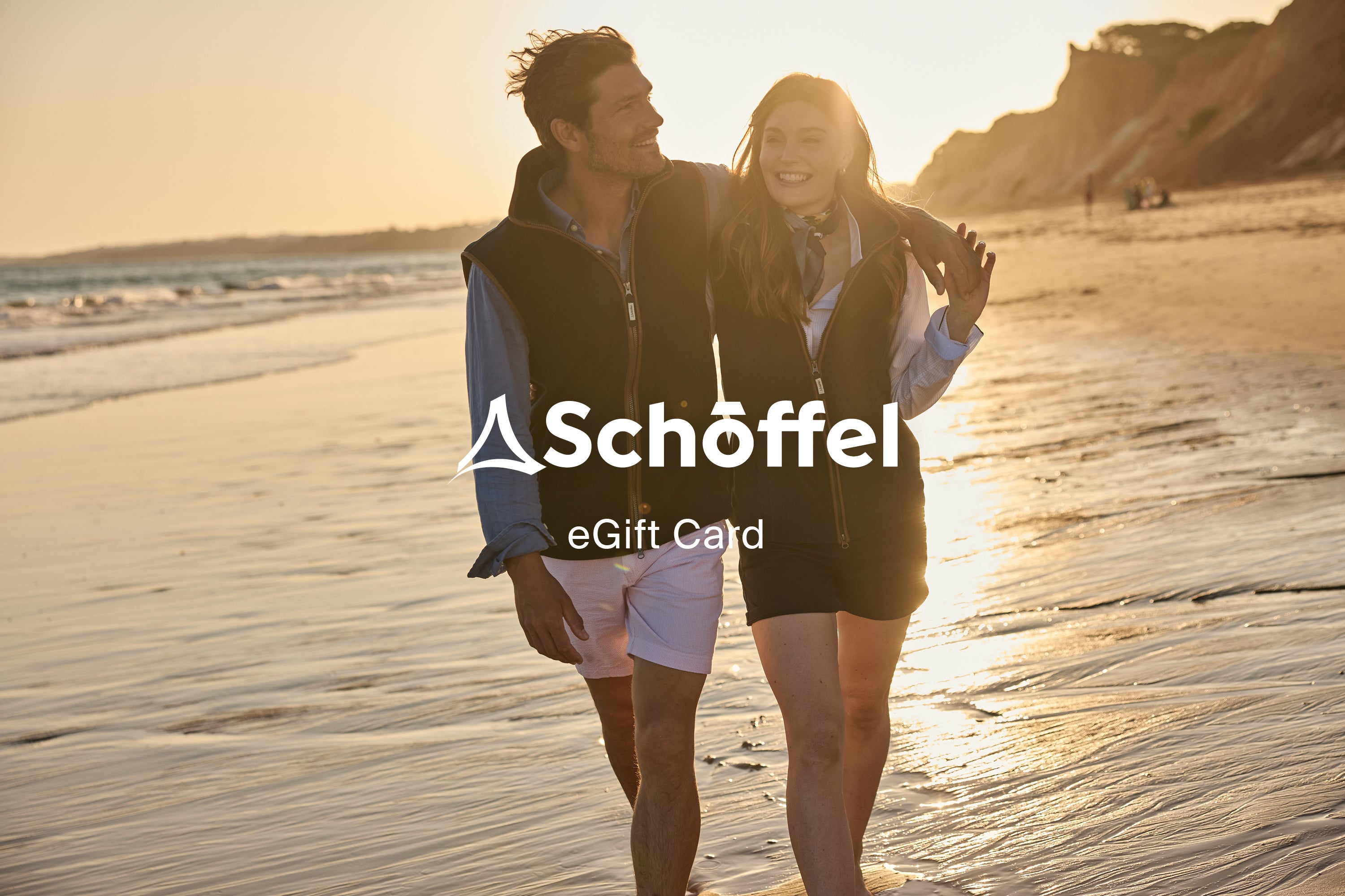 A smiling couple walks along a sandy beach at sunset, with the white text 'Schöffel eGift Card' overlaid across the center. The scene is warmly lit by the setting sun, with ocean waves and cliffs in the background.