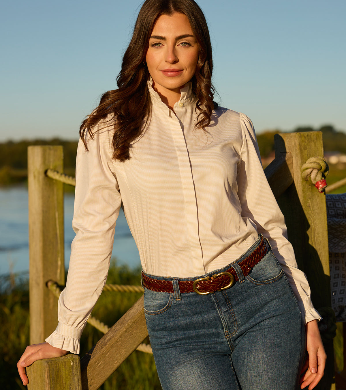 Woman wearing a white shirt and blue jeans standing by a wooden fence with a scenic background.