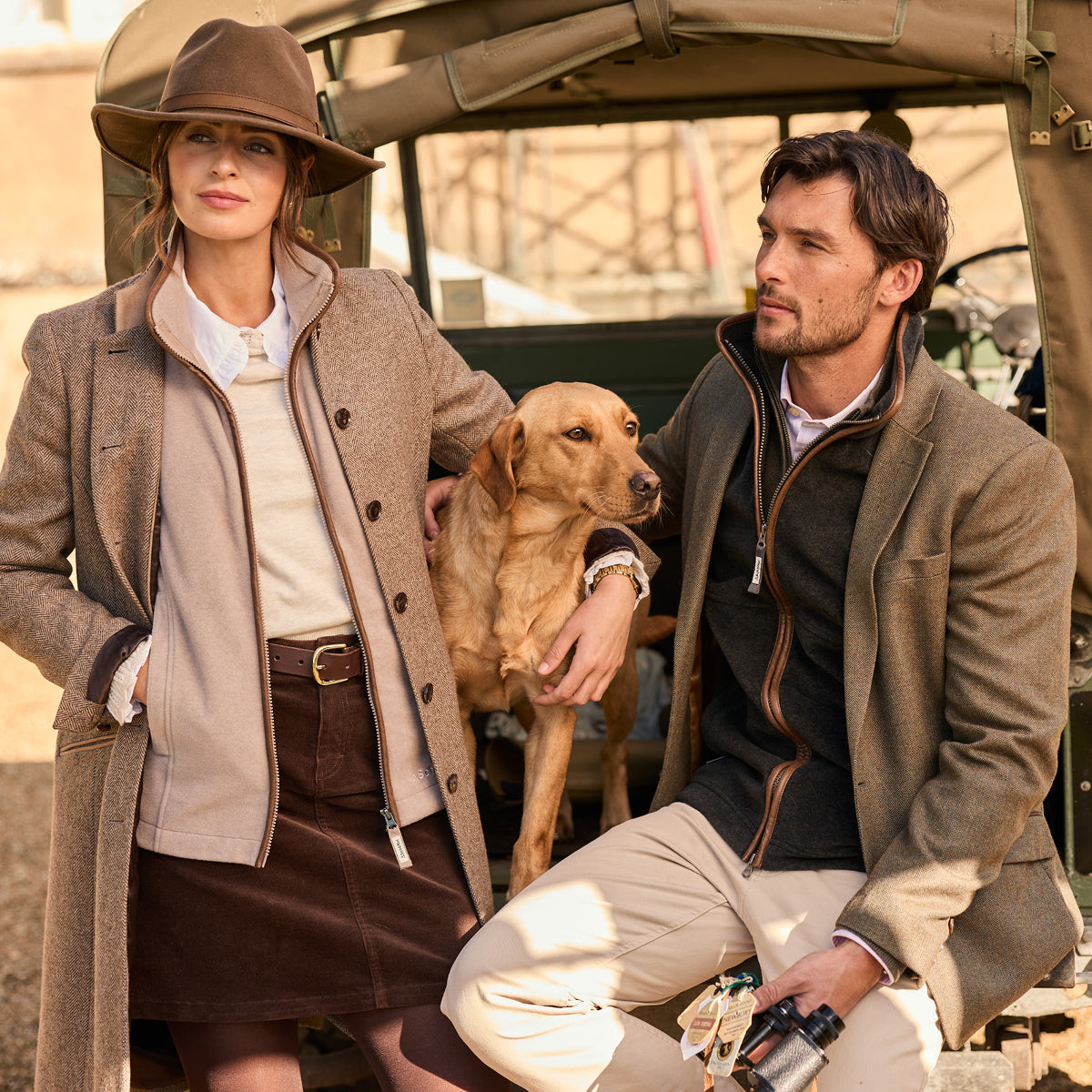 Man and woman with a dog sitting in front of an open vehicle, wearing winter coats.
