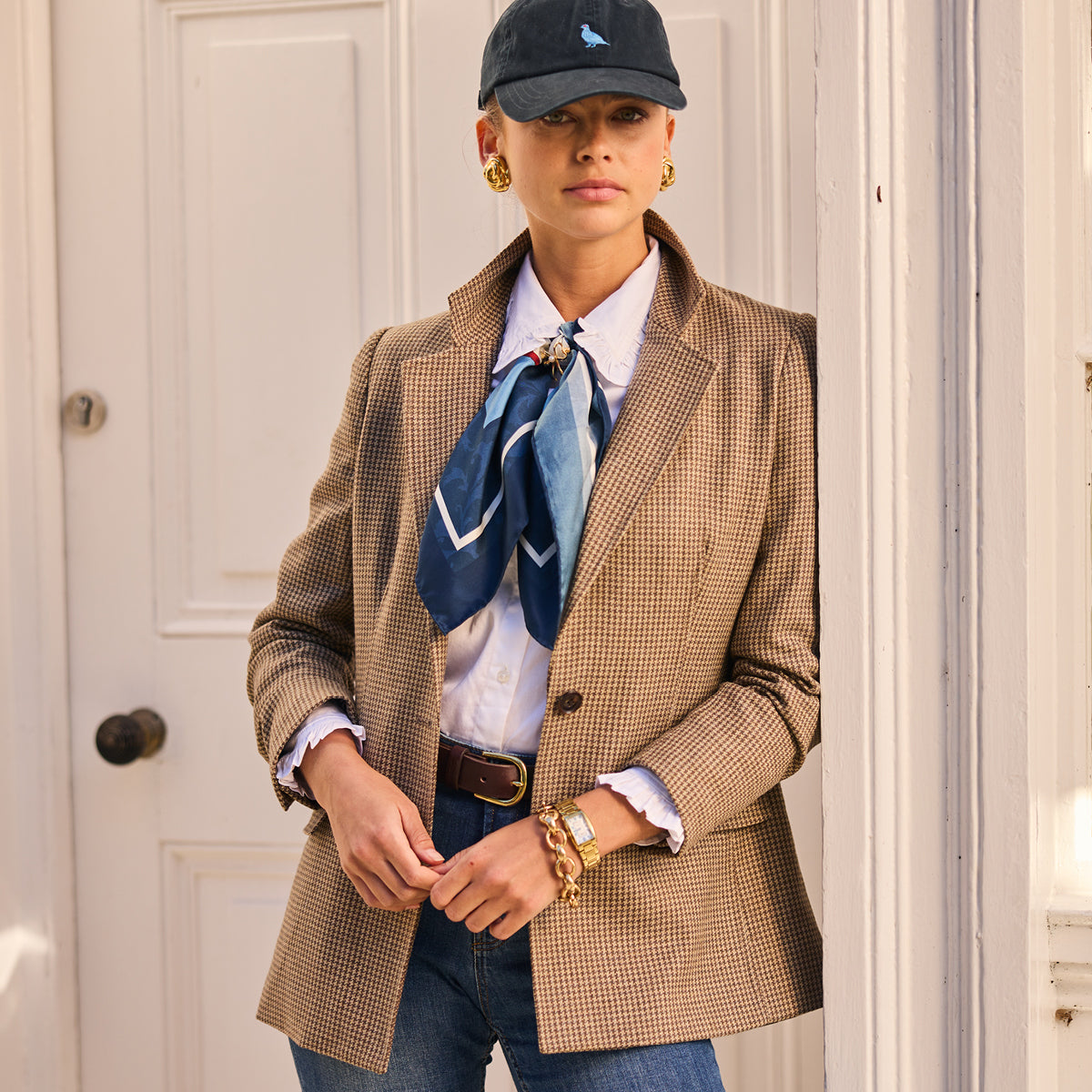 Person wearing a brown tweedblazer with a blue scarf, standing in front of a white door.
