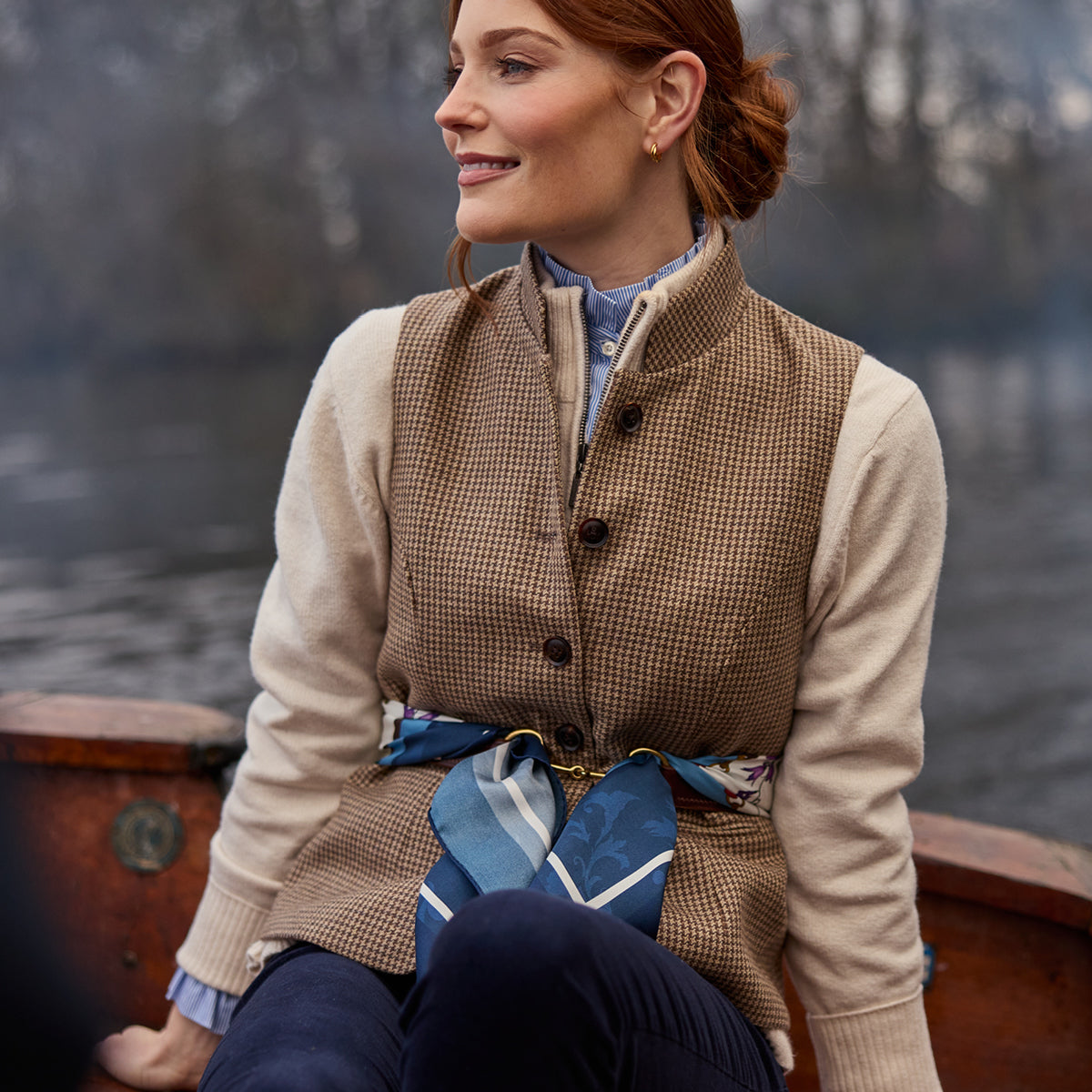 Woman sitting outdoors wearing a patterned vest and scarf, with a blurred natural background.
