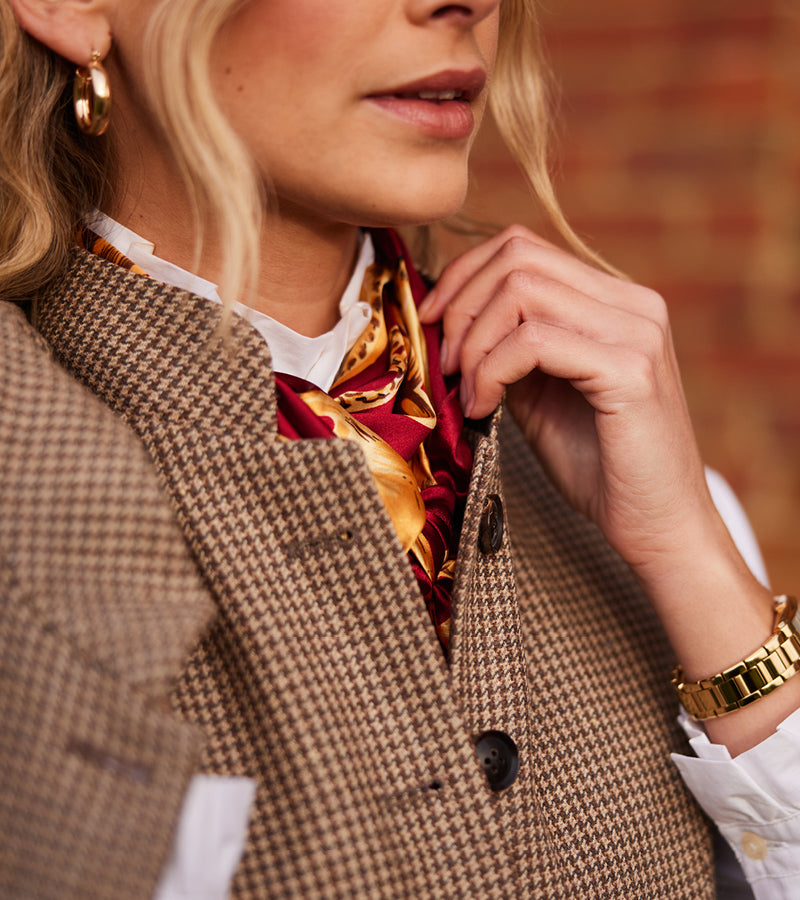 Woman wearing a brown tweed gilet with a colorful scarf and gold watch, against a blurred brick wall background.