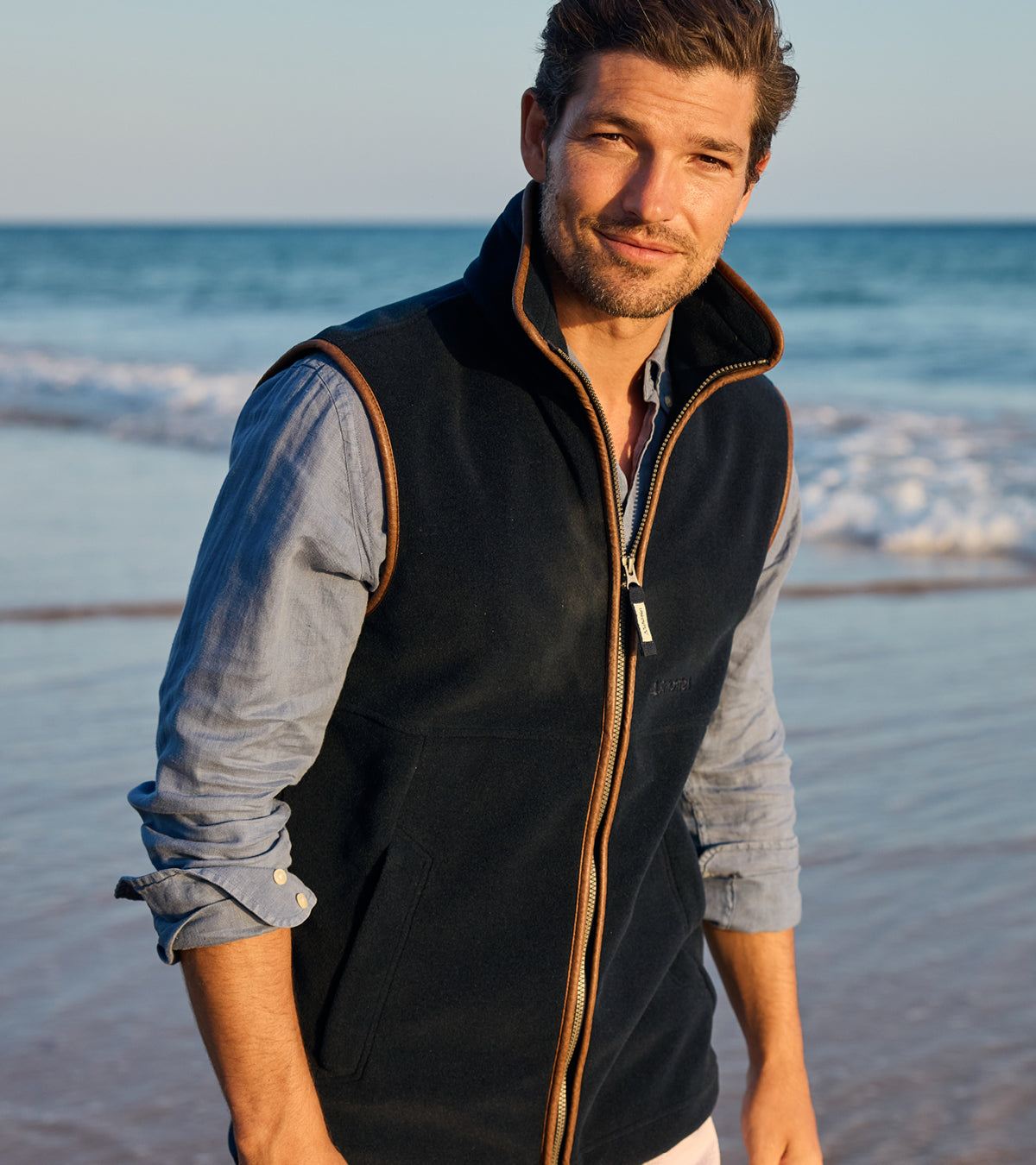 Man wearing a dark gilet over a light blue shirt on a beach