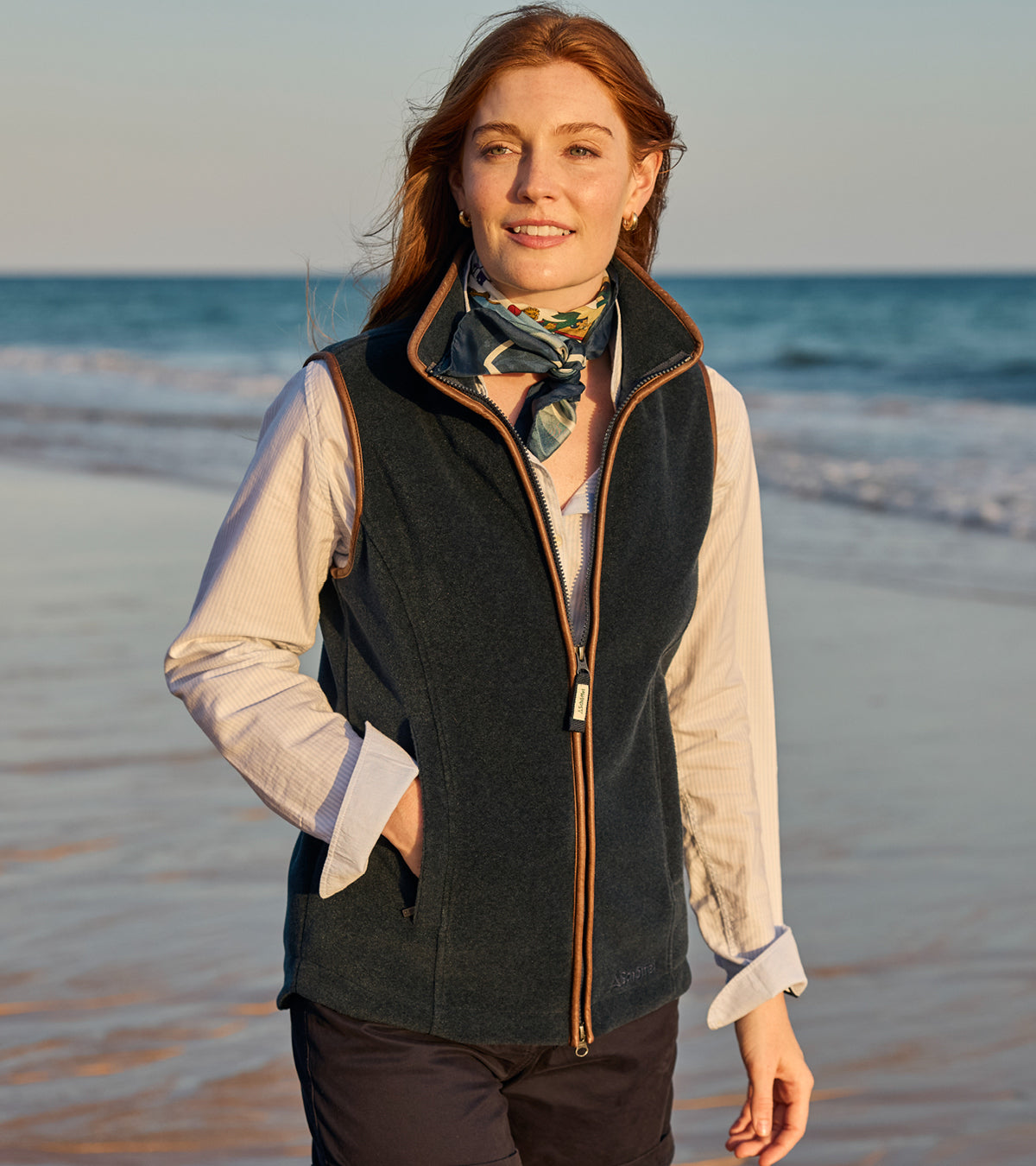 Woman wearing a dark vest and scarf on a beach with ocean in the background