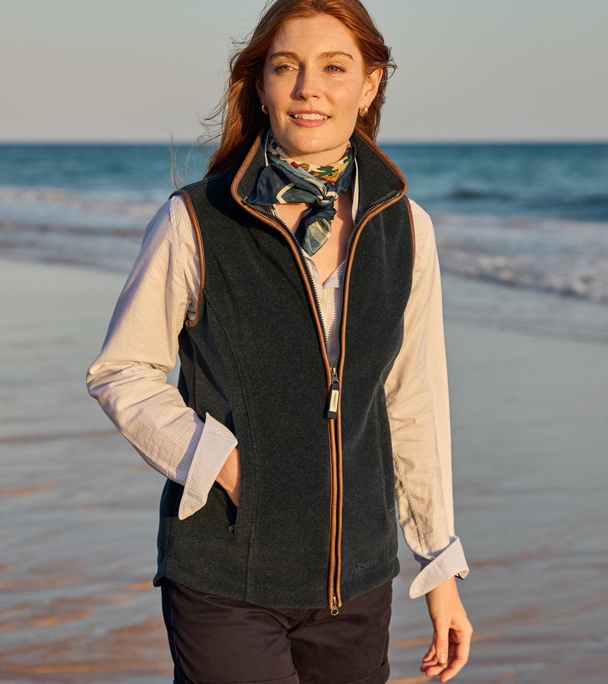 Woman standing on a beach wearing a dark gilet and scarf with ocean waves in the background