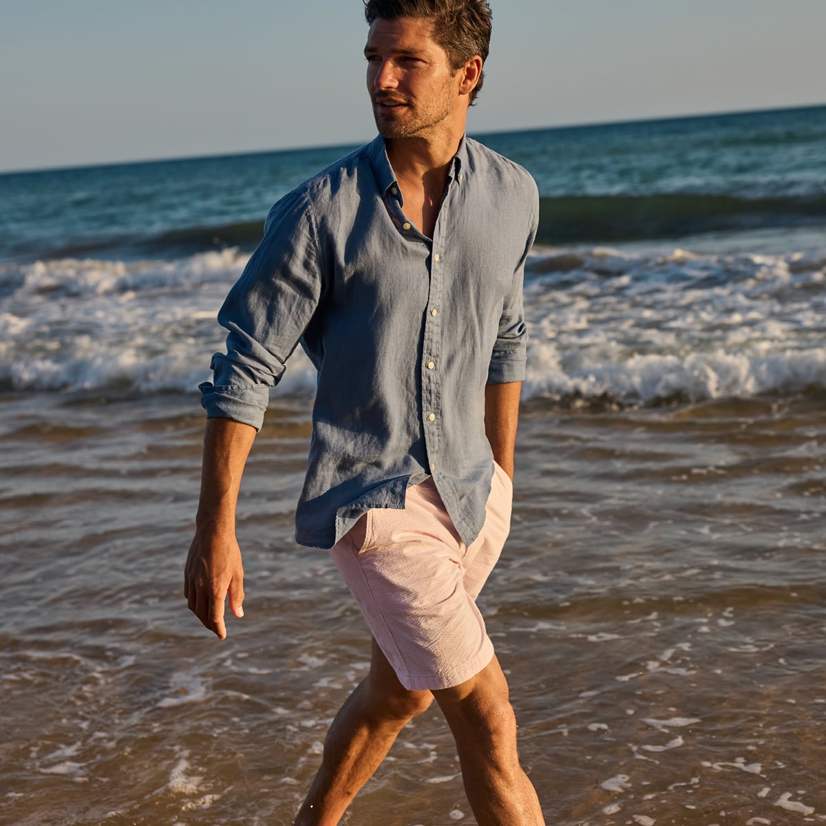 Man walking on a beach with ocean waves in the background wearing a linen blue shirt