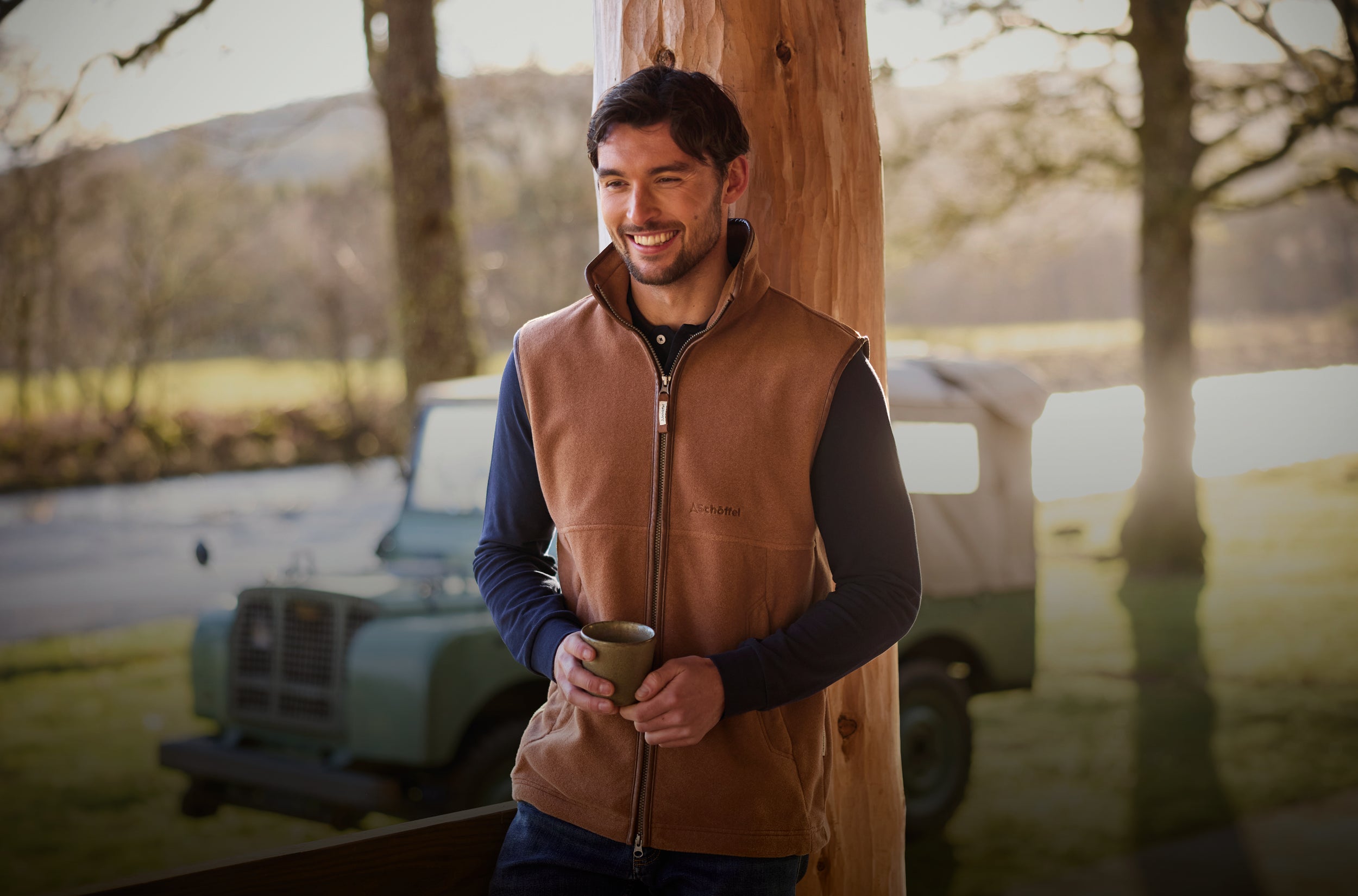 Man wearing a brown gilet holding a mug outdoors with a vehicle and natural scenery in the background