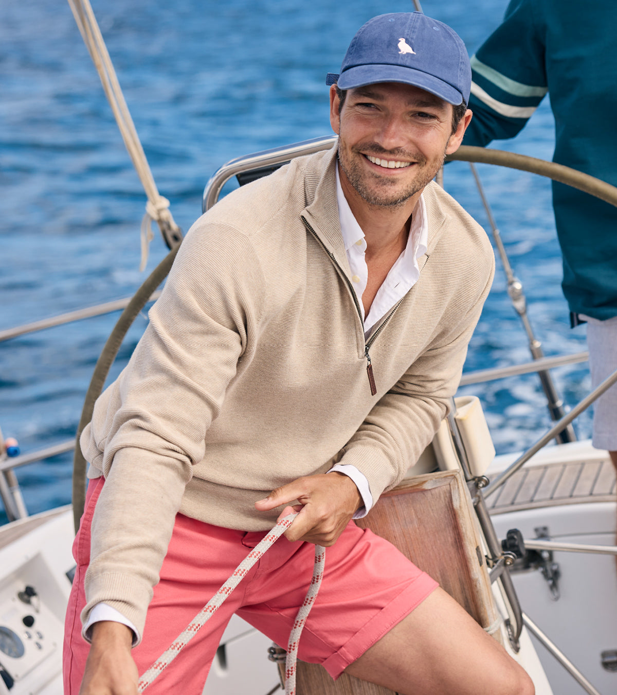 Man on a sailboat wearing a beige sweater, pink shorts, and a blue cap with a logo.