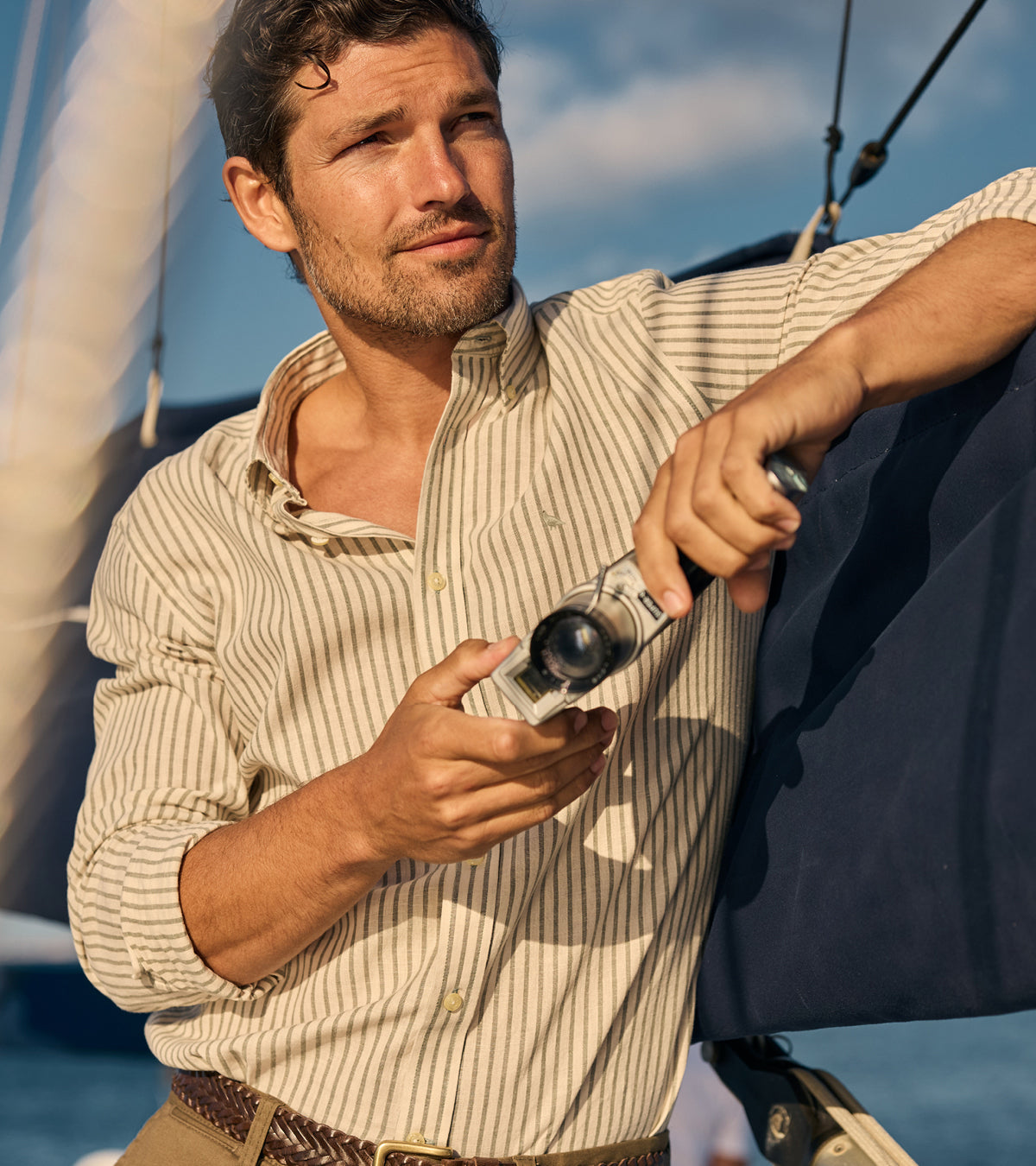 A man on a sailboat wearing a blue and white vertically striped button-down shirt, holding a camera. The background shows blue sky and white sails.