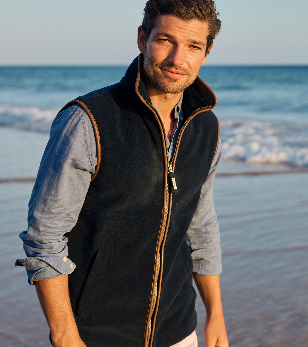 A man standing on a beach wearing a navy zip-up fleece gilet with tan trim over a light blue button-down shirt.