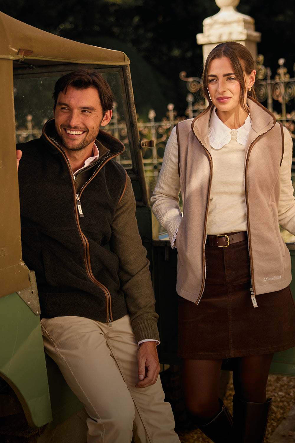 A man and a woman standing outdoors beside a vintage vehicle. The man wears a dark  zip-up fleece gilet and beige trousers, while the woman wears a beige zip-up gilet over a cream blouse and a brown skirt.