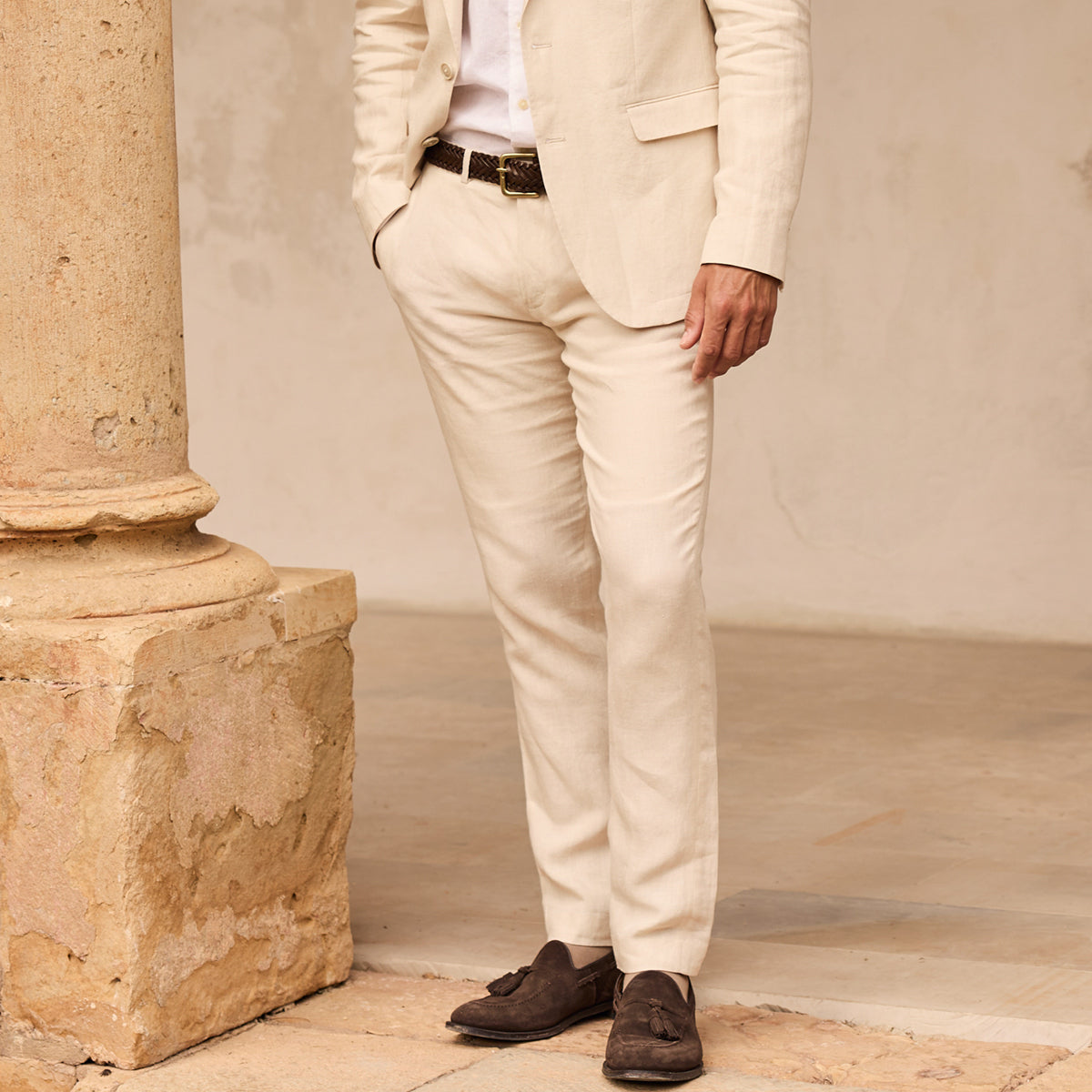A man wearing a light beige linen suit, including a blazer and trousers, styled with a white shirt, a dark brown belt, and dark brown suede tassel loafers, standing next to a stone column.