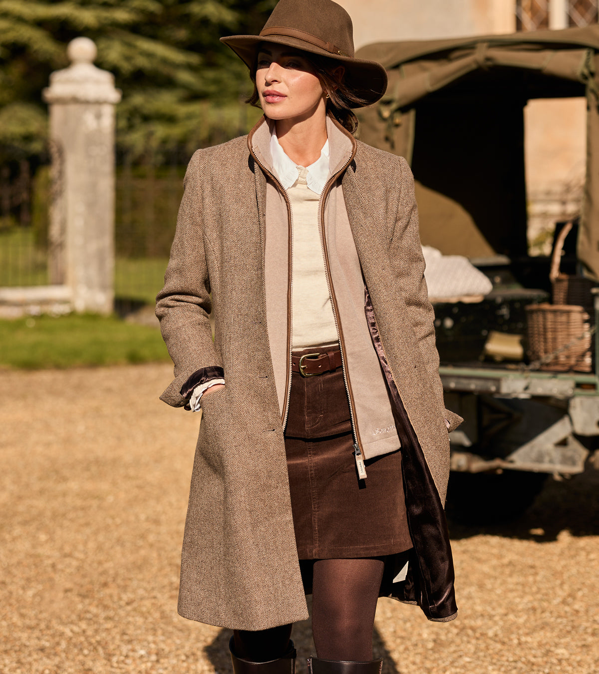 Woman in a brown coat and hat standing outdoors with a vintage car in the background