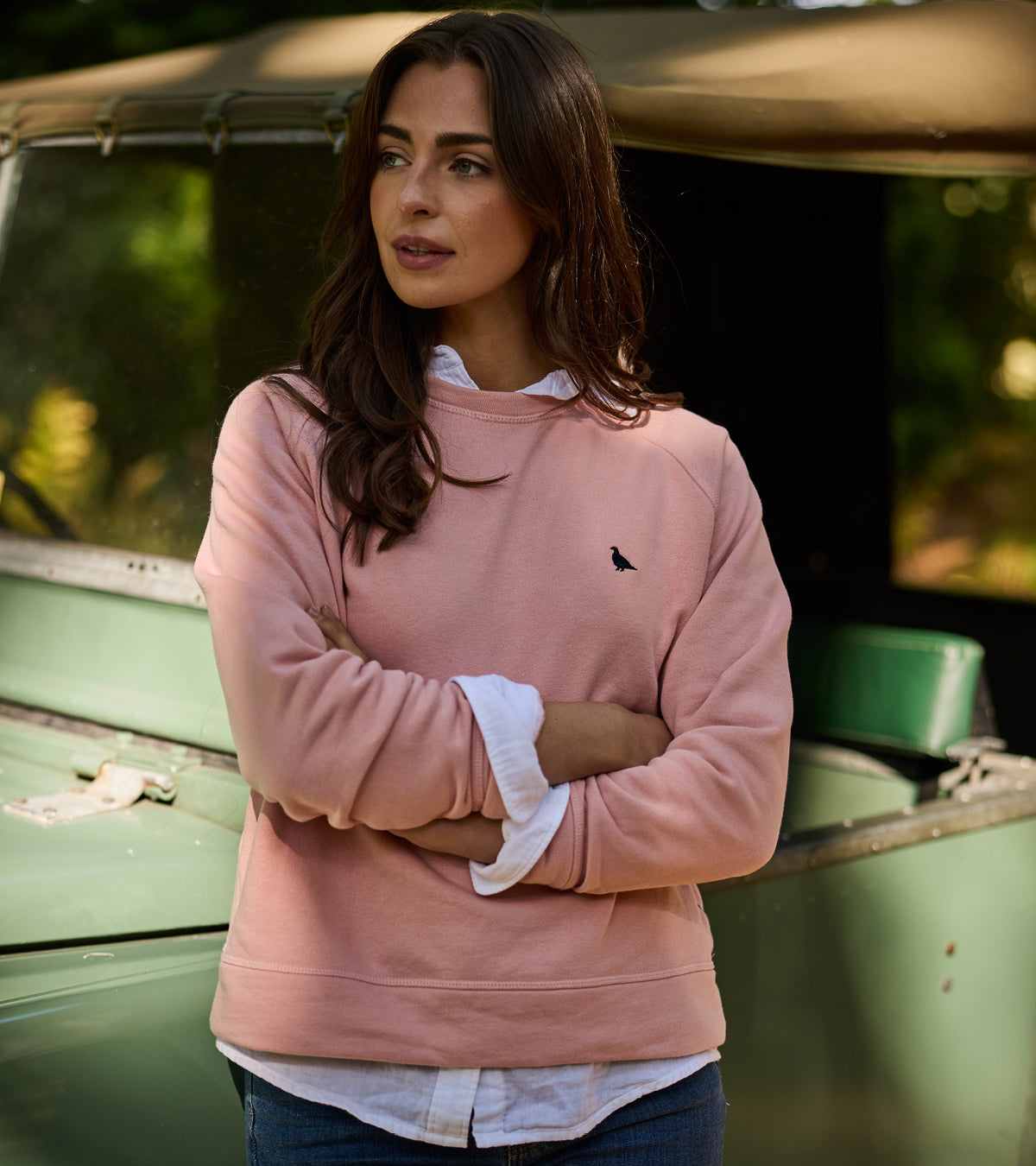 Woman wearing a pink sweatshirt with a small logo, standing in front of a vintage vehicle.