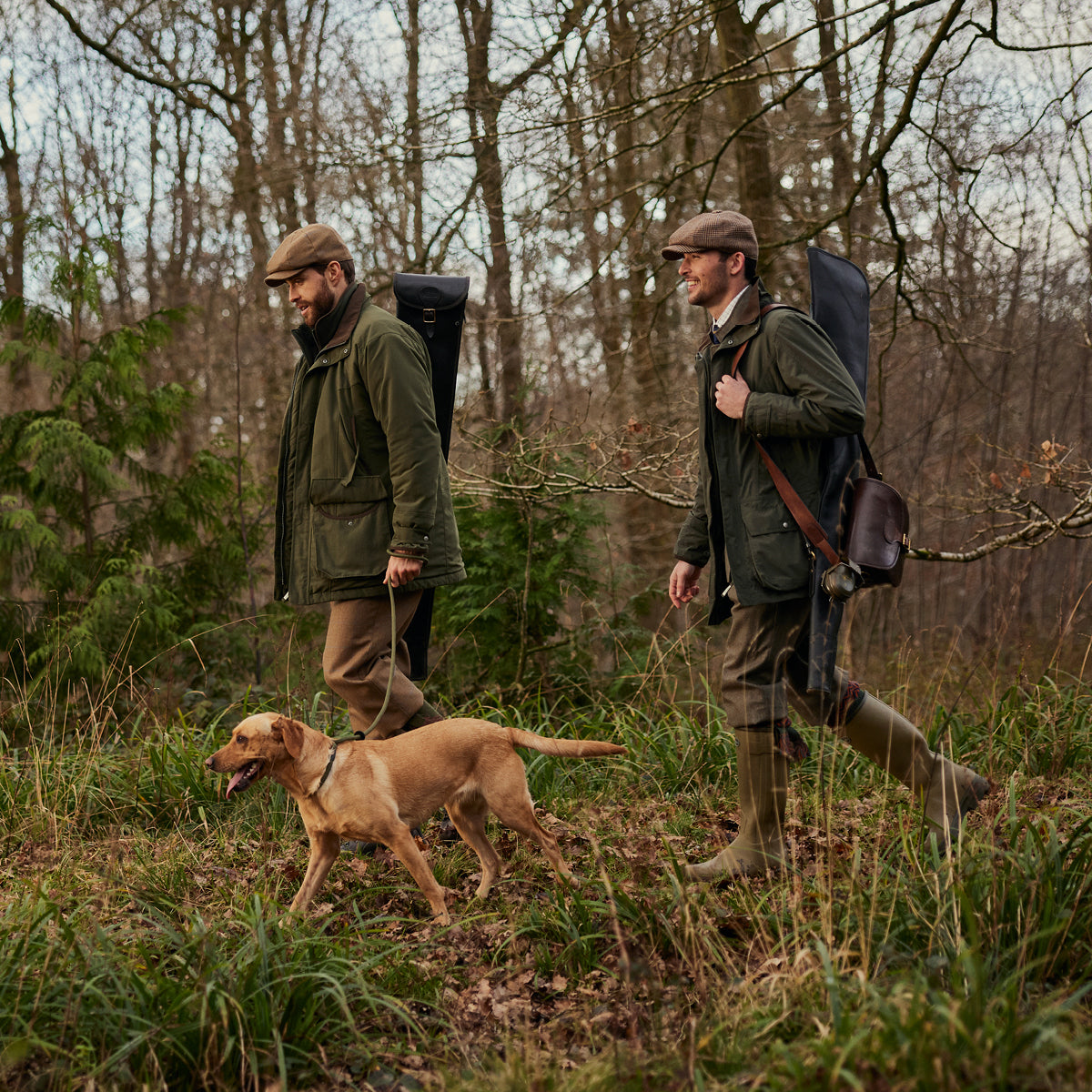 An image of two men and dog walking through a woodland area in countrywear.