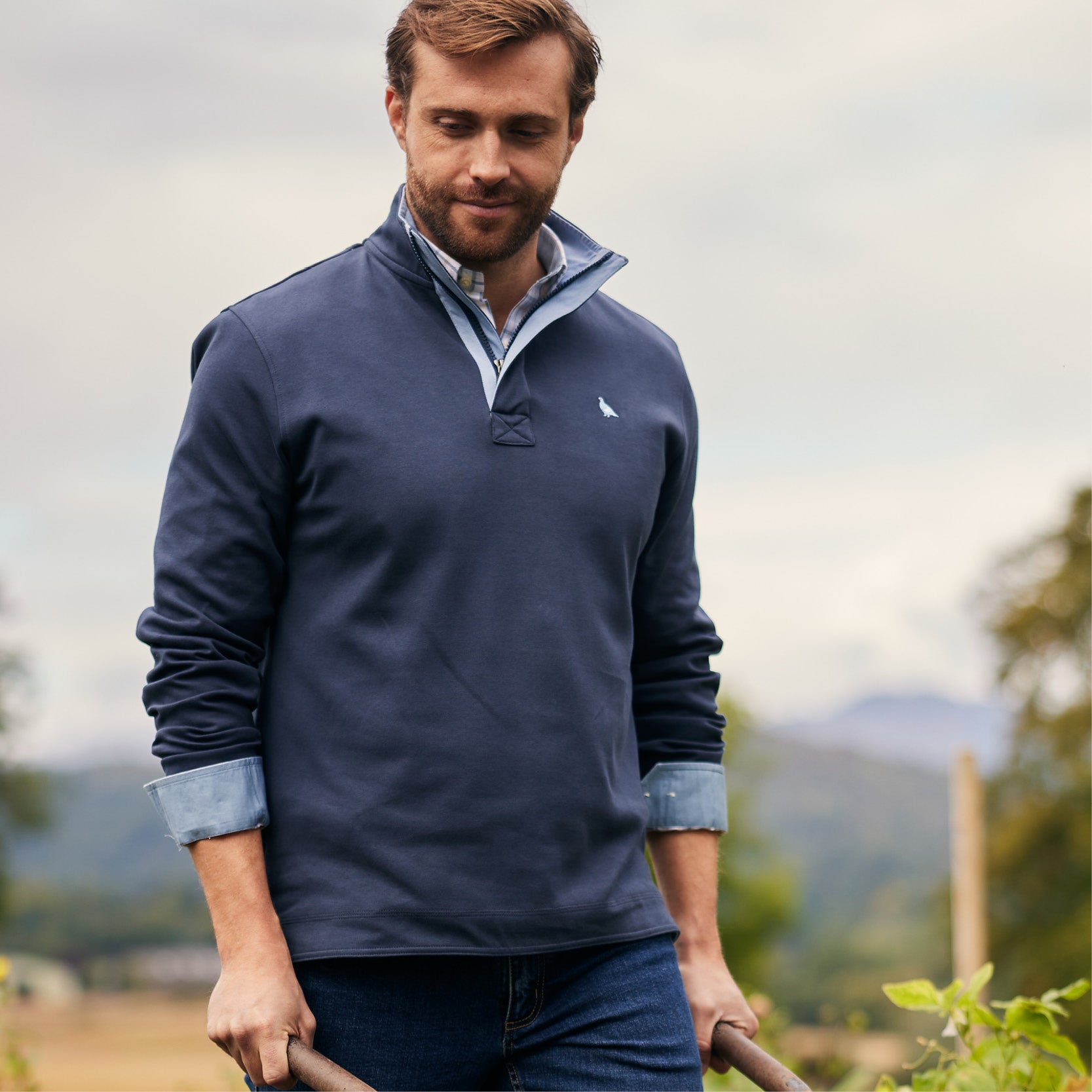 An image of a man wearing a blue sweatshirt with a checked shirt underneath, he is wearing the cuffs rolled up and is pushing a wheelbarrow in a garden area.