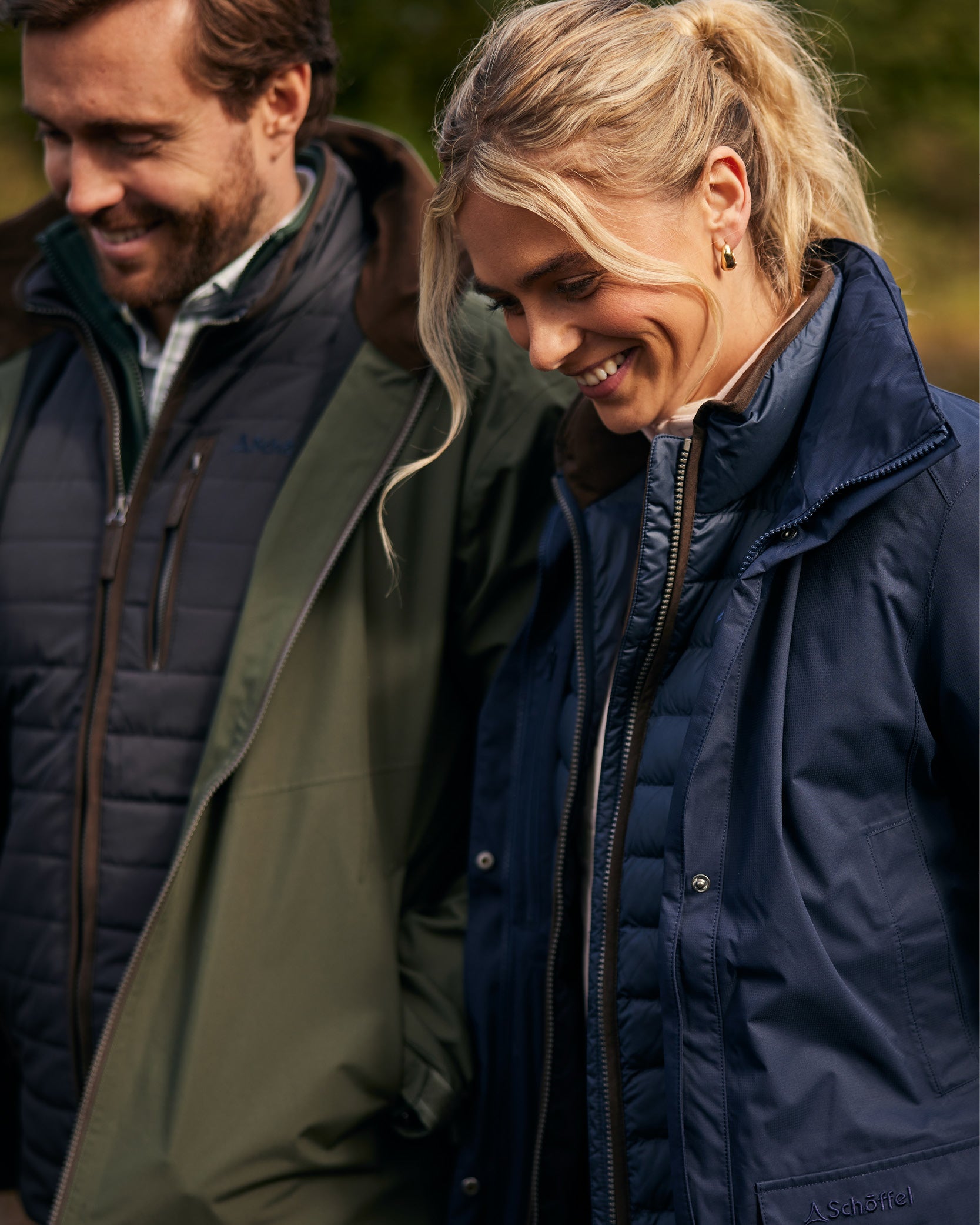 A man and a women are smiling walking in an outdoor setting. He is wearing a blue gilet and green jacket layered over a checkered shirt, she is wearing a navy gilet and jacket layered over a light pink sweatshirt.