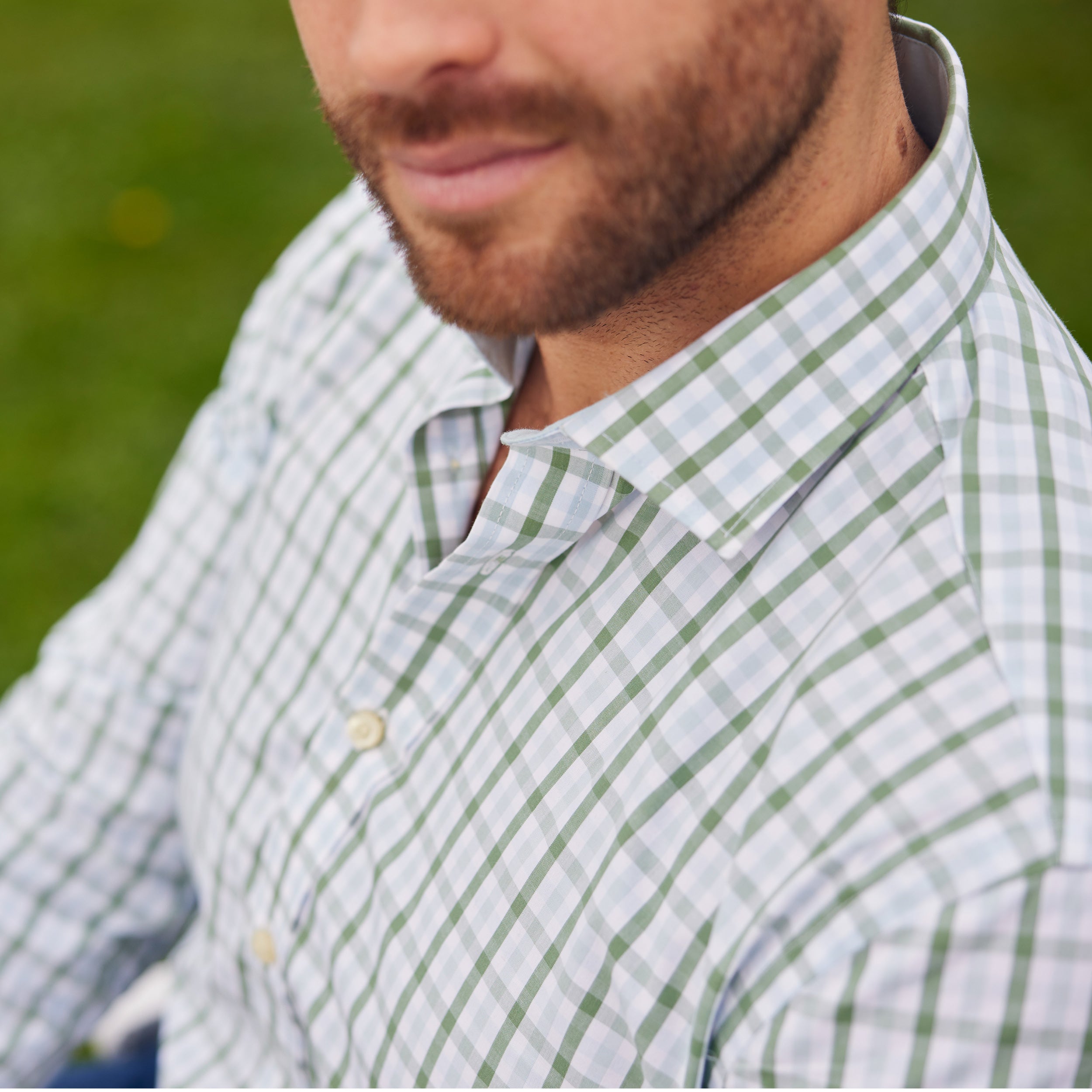 A close-up shot shows a man's face from the nose down. He has a short, well-groomed beard and wears a light-coloured button-down shirt with a green and white plaid pattern. The collar of the shirt is visible, and the background is a blurred green.