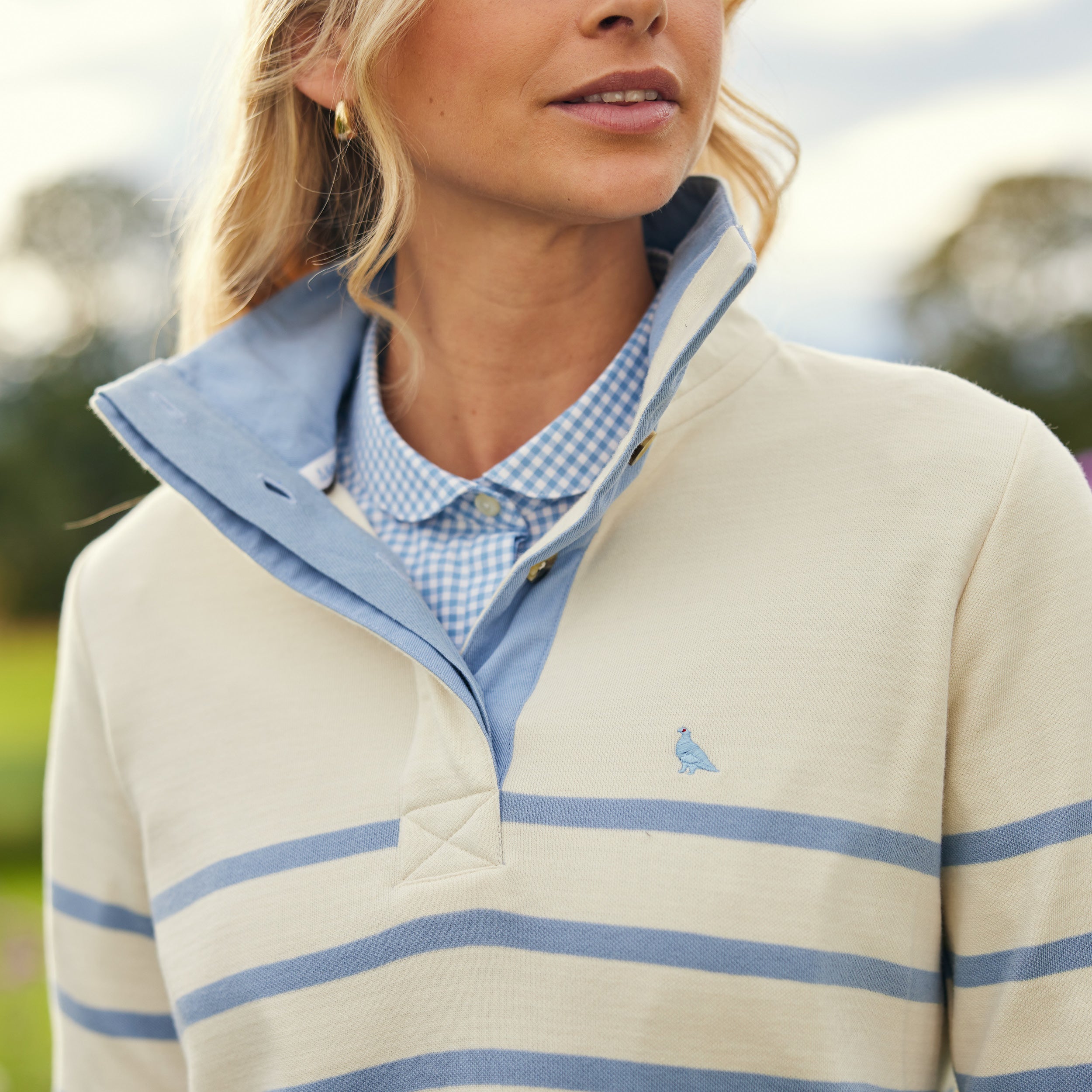 A woman with blonde hair and gold hoop earrings wears a cream-colored, horizontally striped sweatshirt with blue accents over a light blue and white gingham collared shirt