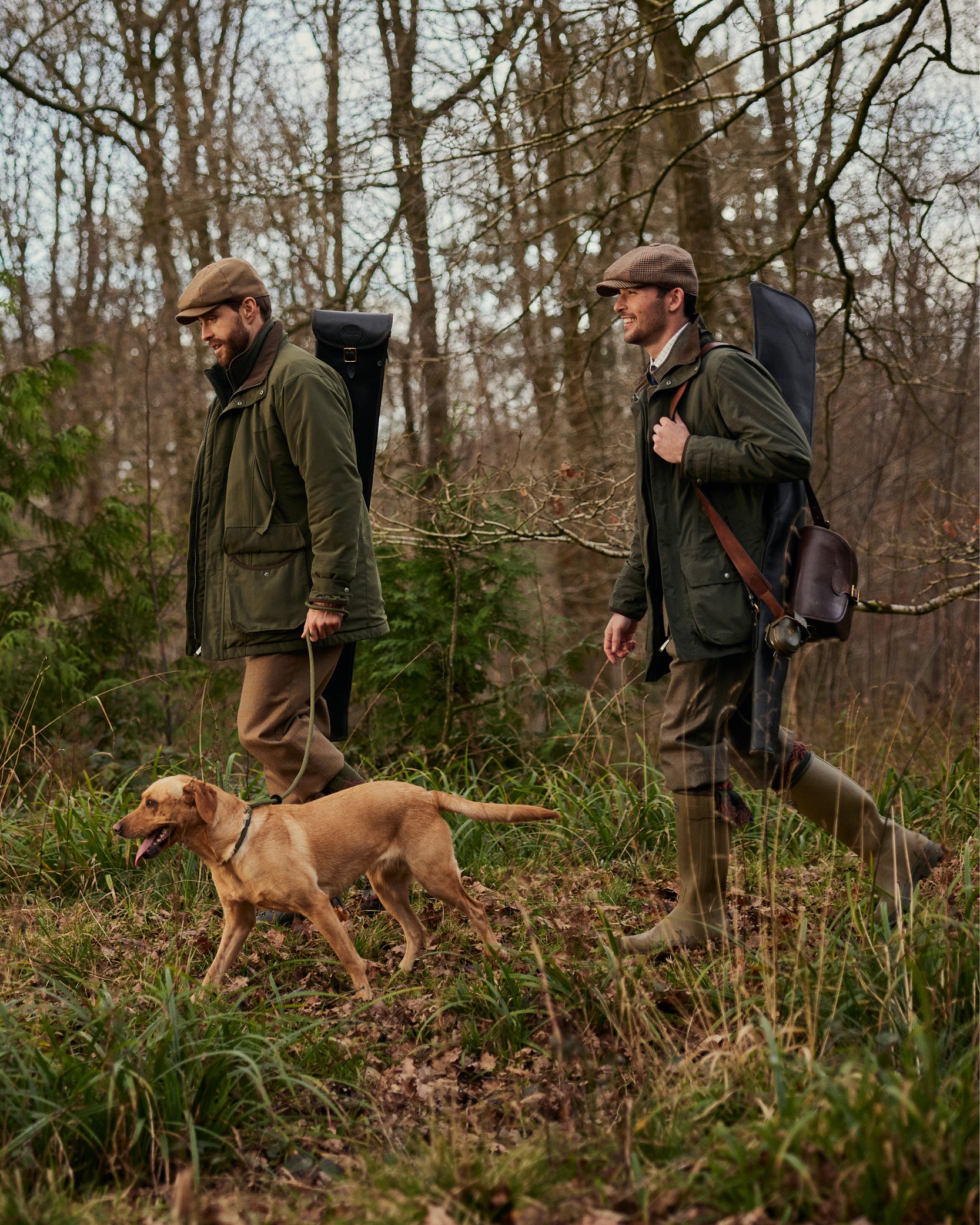 Two men in outdoor attire, including tweed caps and green jackets, walk through a wooded area. One man has a dog on a lead,  both carry gun cases over their shoulders.
