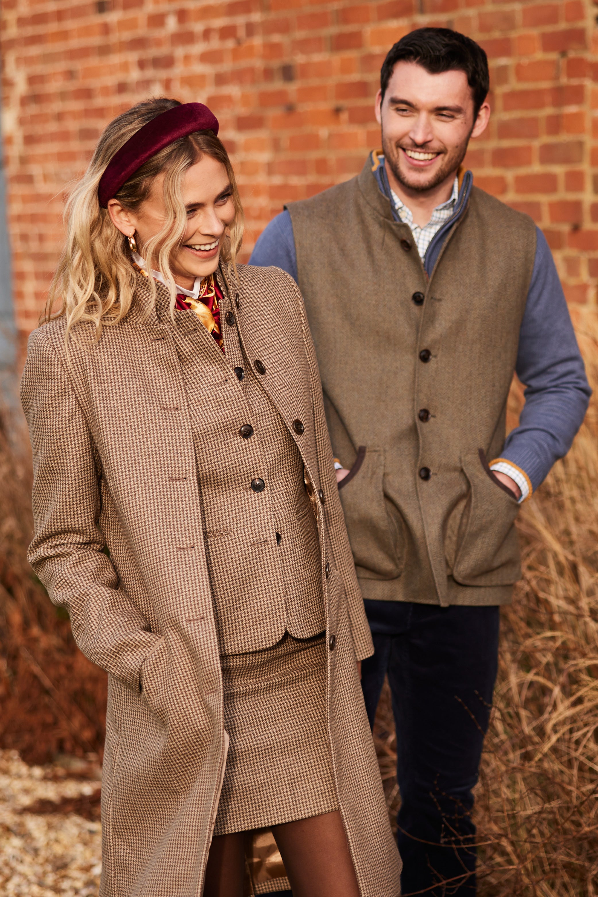 A smiling man and woman stand outdoors near a brick wall and tall, dry grass. The woman has blonde hair, wears a brown tweed coat and skirt, and a burgundy headband. The man has dark hair, wears a brown tweed vest over a blue long-sleeved shirt, and dark trousers