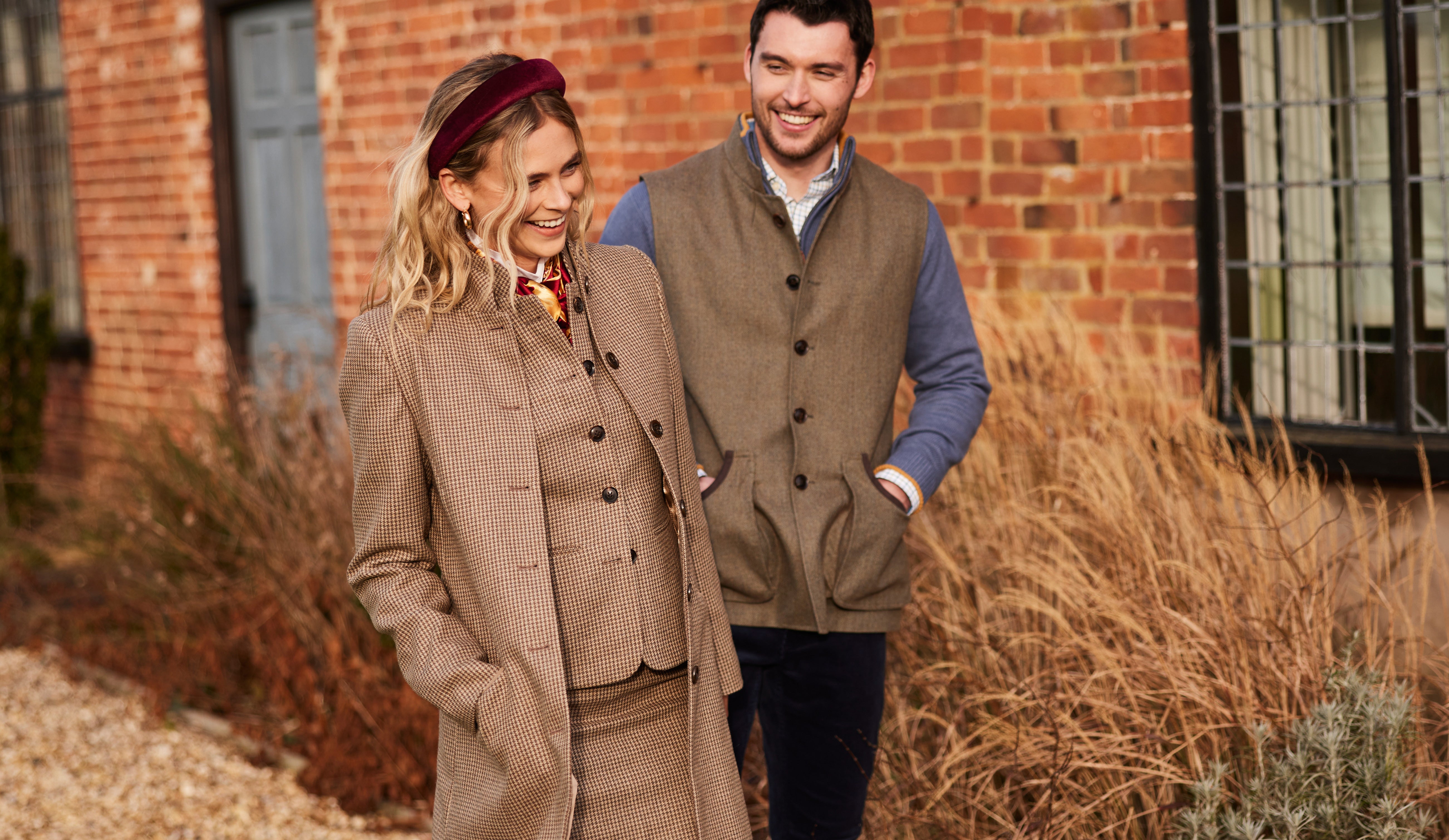 A smiling man and woman stand outdoors near a brick wall and tall, dry grass. The woman has blonde hair, wears a brown tweed coat and skirt, and a burgundy headband. The man has dark hair, wears a brown tweed vest over a blue long-sleeved shirt, and dark trousers