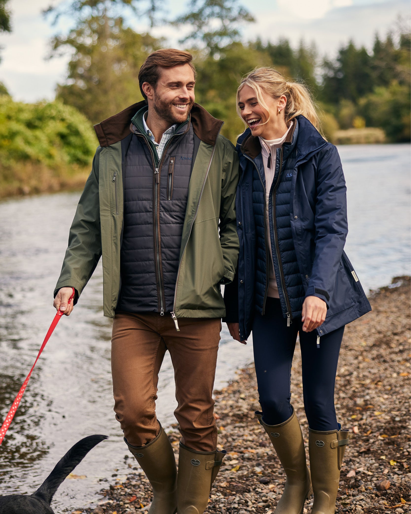 An image of a couple walking arm in arm along a waters edge. They are wearing country outerwear clothing and leading a black dog.