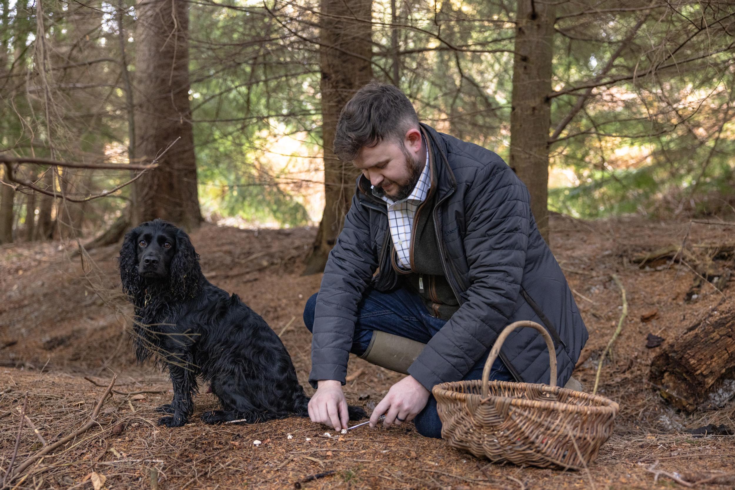 A man is crouching in a woodland area he is with his black dog and has a wicker basket.