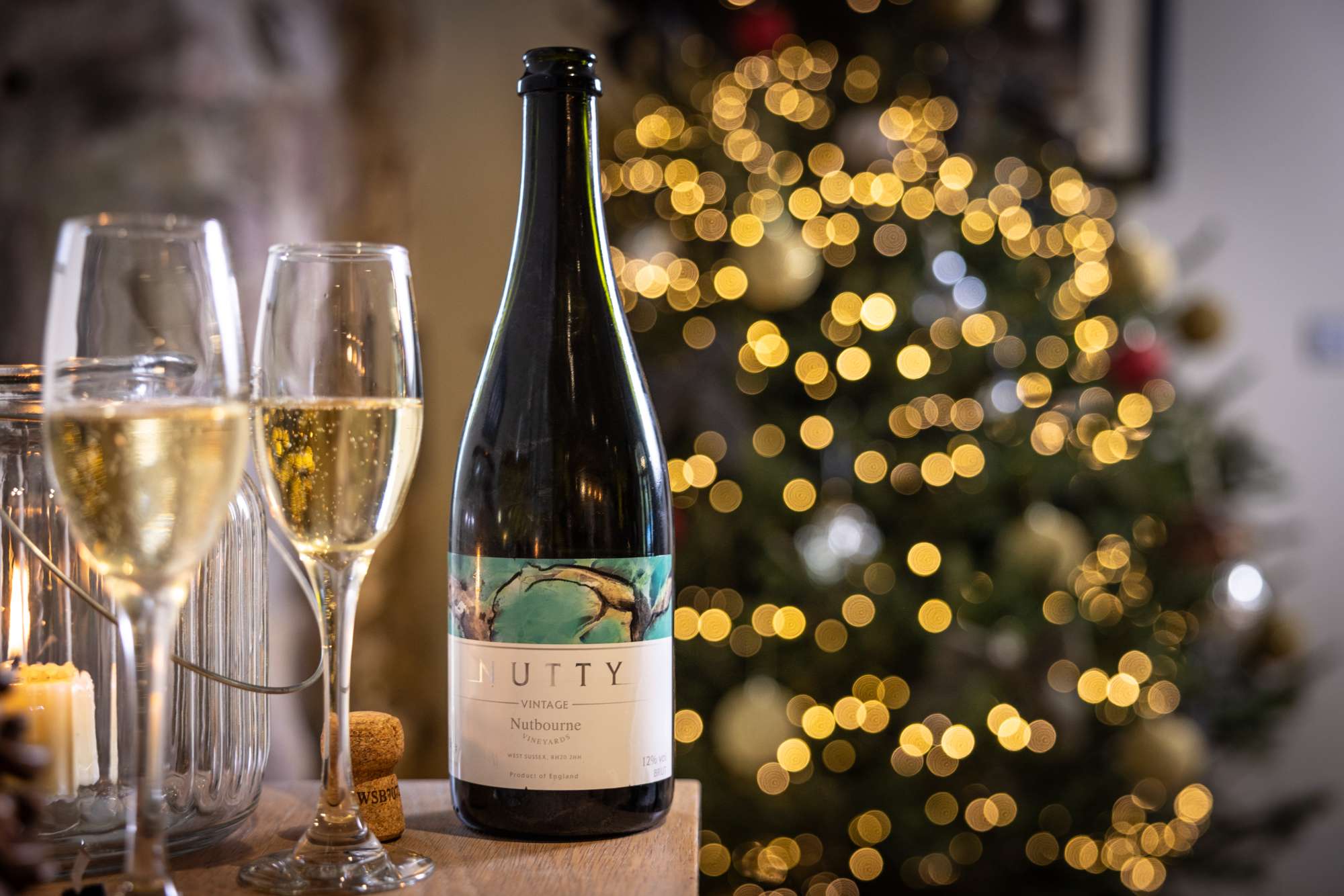 A close up of a bottle and glasses with a Christmas tree in the background.