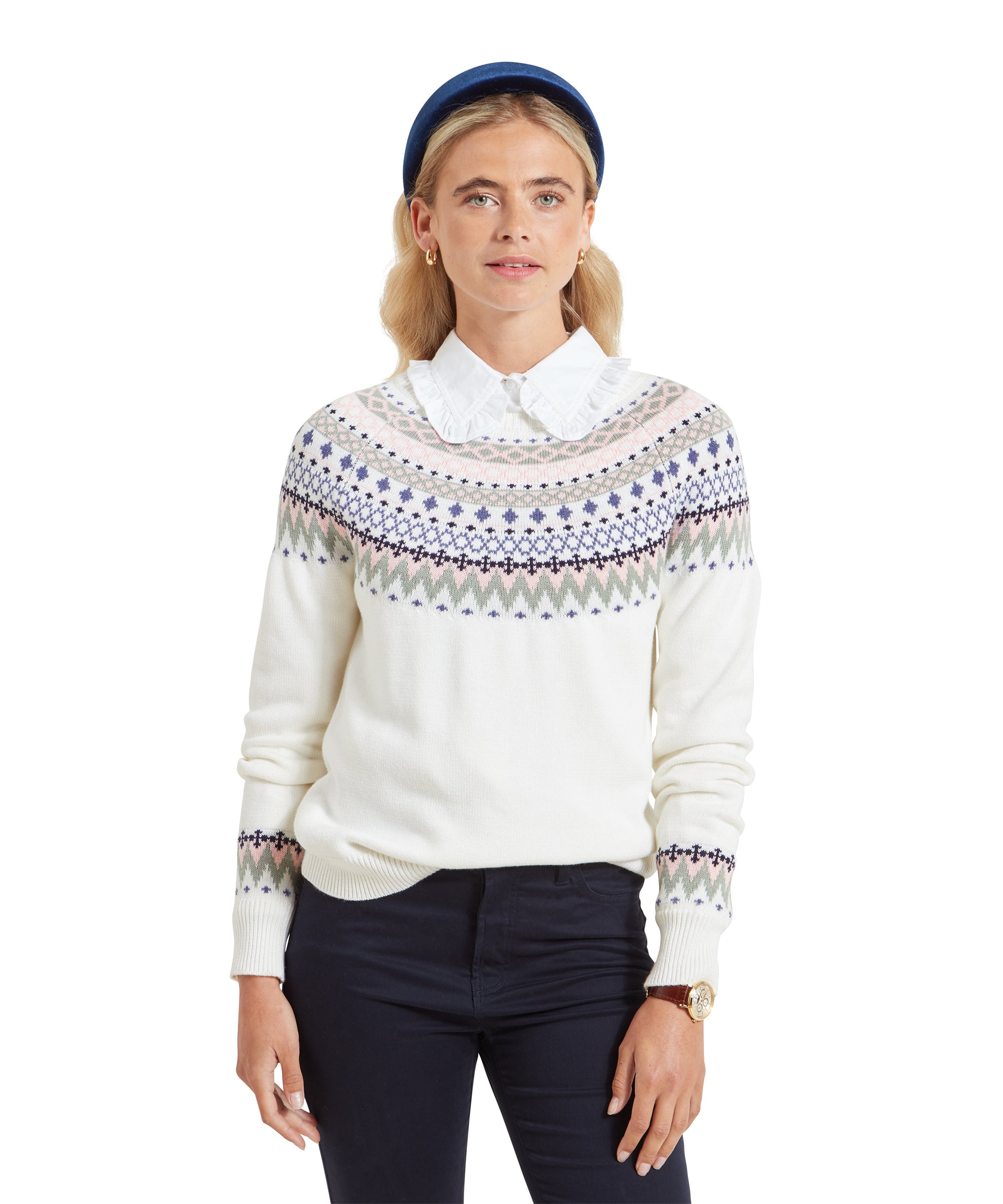 An image of a lady wearing a white fairisle knitted jumper with a white shirt and navy trousers. She is wearing gold accessories and a navy headband and she is looking towards the camera.