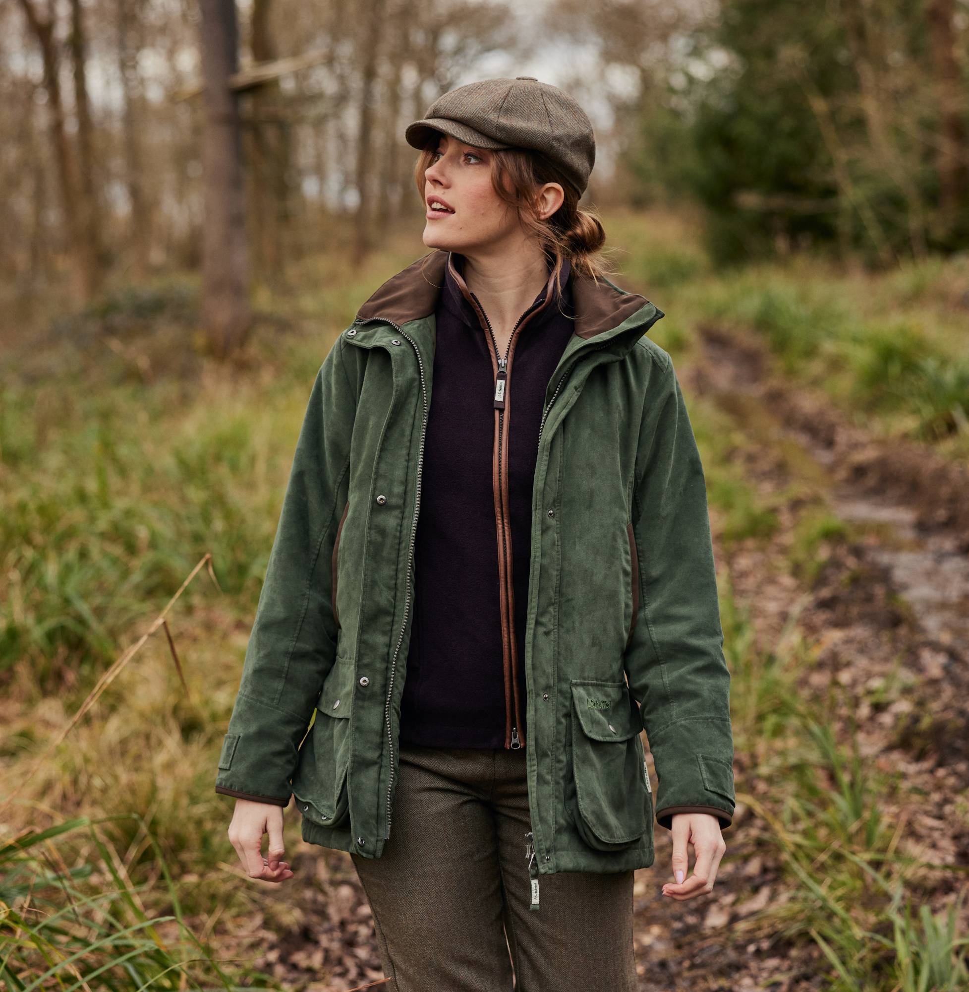 A woman wearing a dark purple fleece, tweed trousers and cap and a country green coat. She is in woodland and she is smiling looking away from the camera.