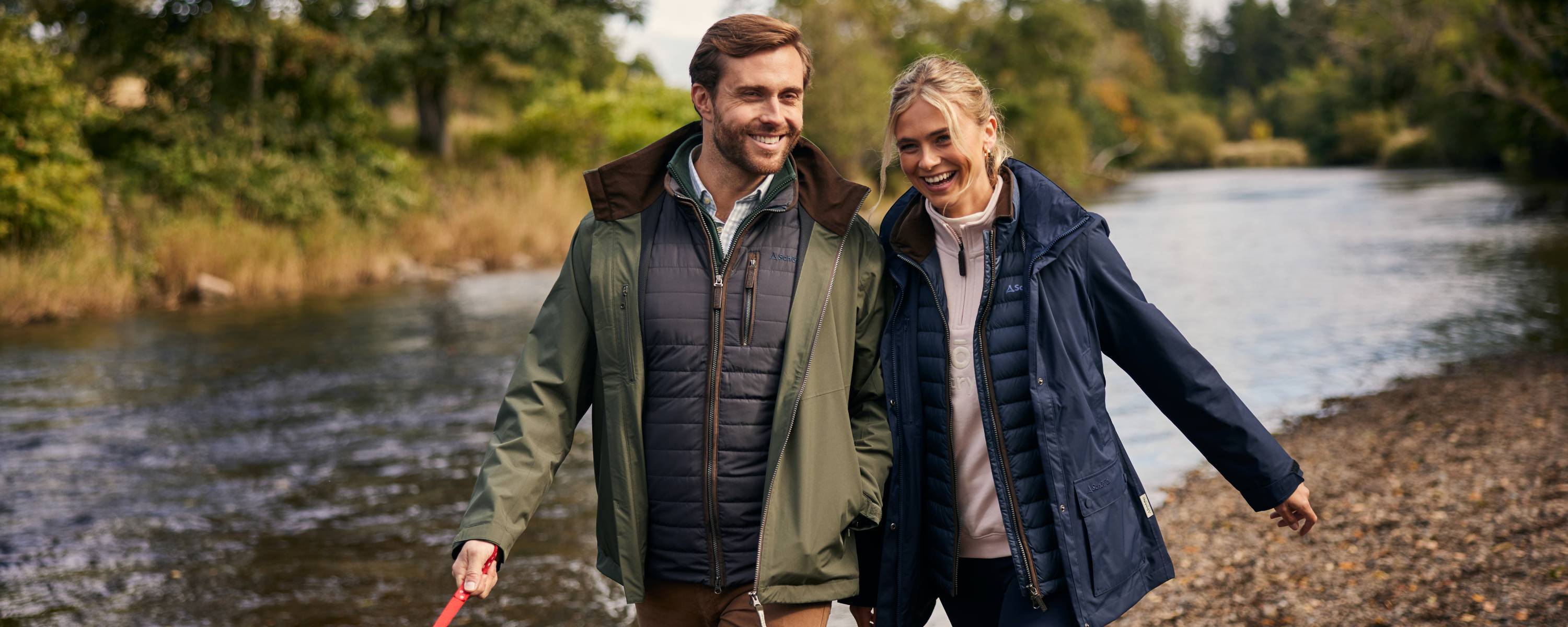 A smiling man and woman walk along the edge of a scenic river, dressed in stylish outdoor clothing. The man is wearing a green waterproof jacket over a navy quilted vest and a checkered shirt, while the woman wears a navy raincoat over a light-coloured sweatshirt. The background features lush green trees and a gently flowing river.
